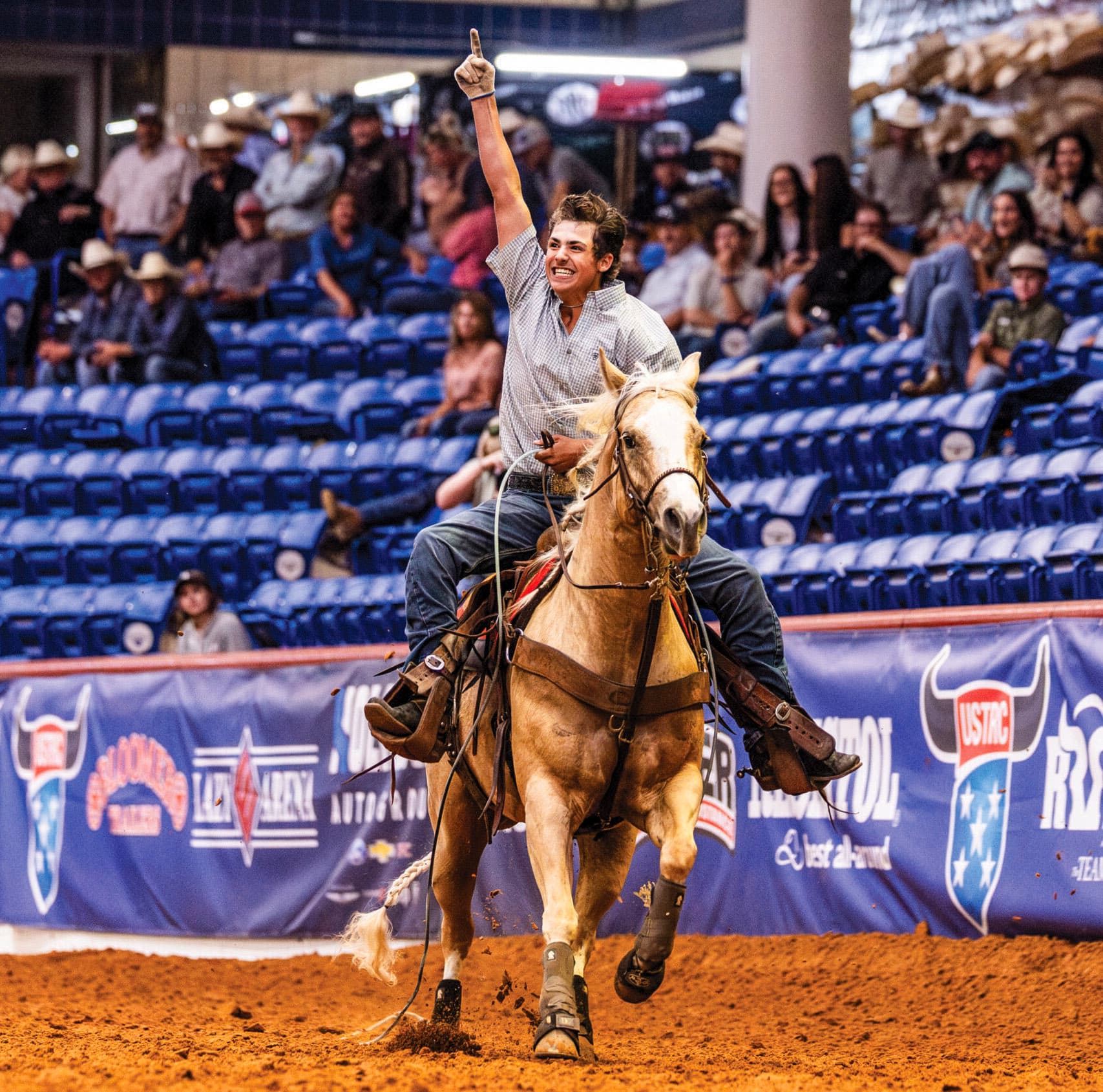 Cowboy on palomino horse raises fist in victory lap around indoor rodeo arena with spectators in blue seats