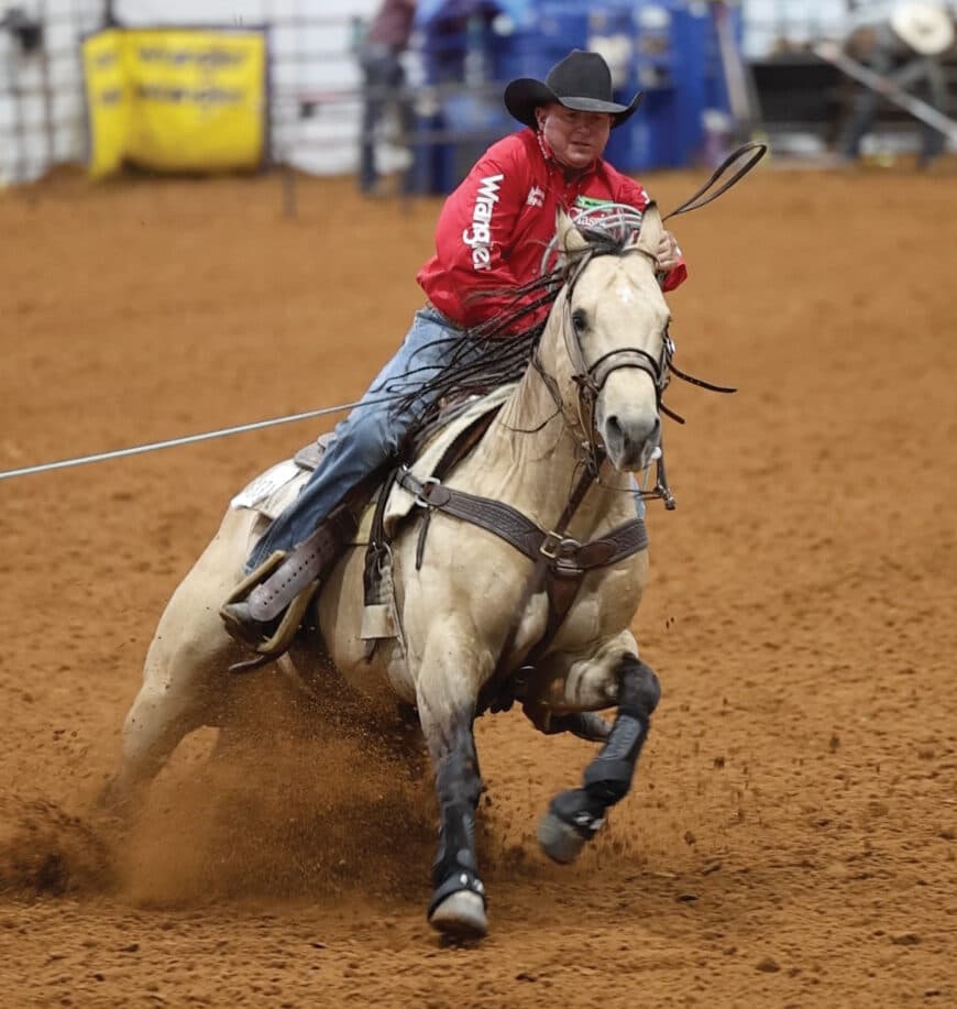Roper on white horse in red shirt and cowboy hat gallops across dirt arena while handling rope during rodeo competition