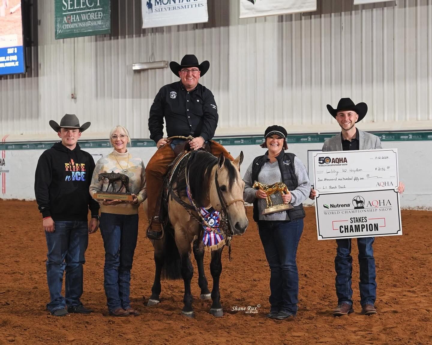 Rider on a buckskin horse flanked by four people holding trophies and an oversized champion check in an indoor arena