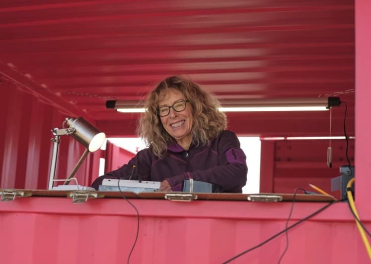 Smiling woman with curly hair and glasses leans over equipment in a pink announcer's booth above the arena