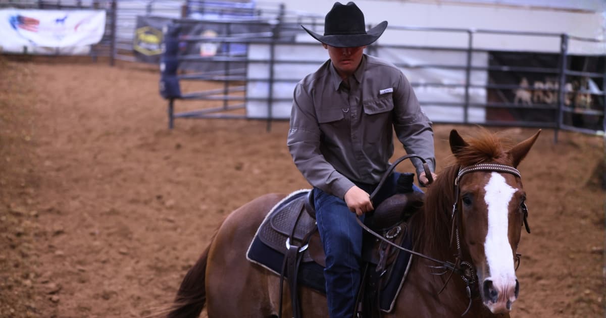 Cowboy in a black hat and gray shirt rides a sorrel horse with a blaze through a dirt arena