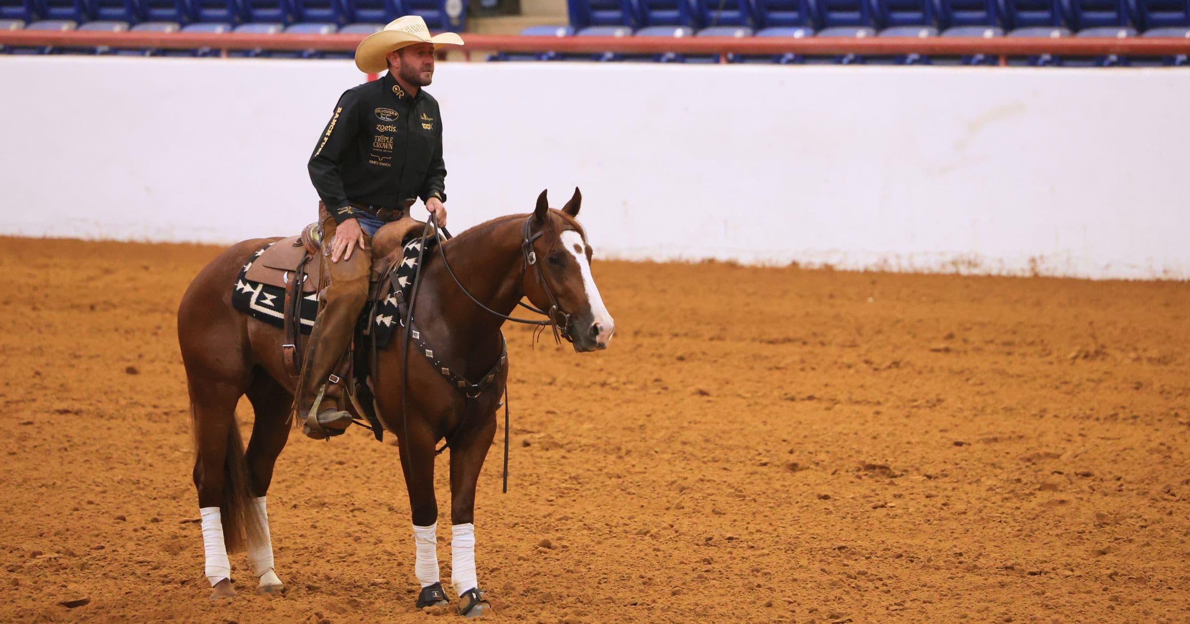 Cowboy in straw hat and black sponsor shirt sits atop a chestnut horse with a blaze, halted in an indoor arena