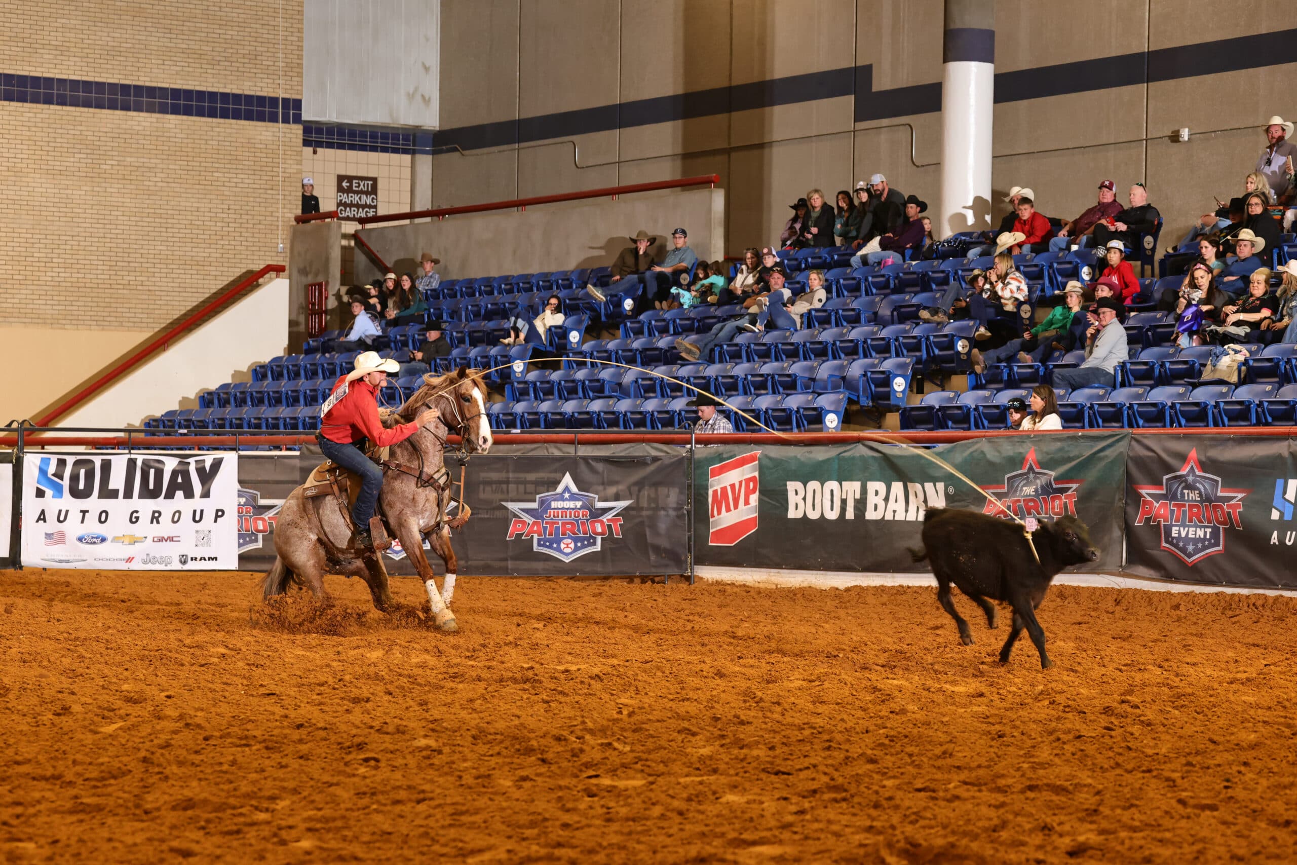 Breakaway roper in a red shirt and white hat rides a roan horse as her loop stretches toward a running calf in the arena