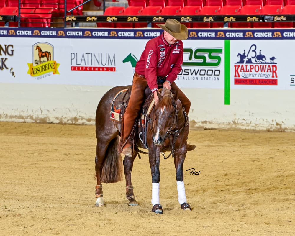 Rider in a cowboy hat and maroon shirt pats the neck of his sorrel reining horse in an indoor arena