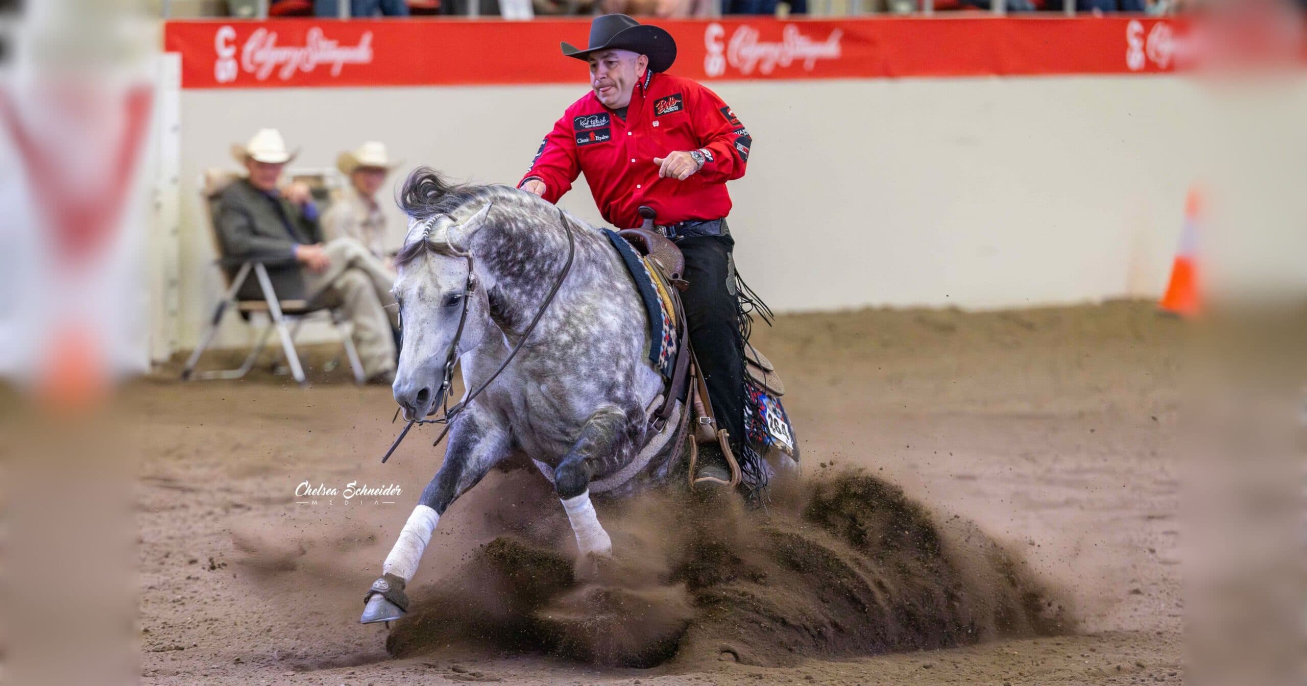 Reiner in a red shirt and black cowboy hat slides his dapple grey horse to a stop, spraying dirt in an indoor arena