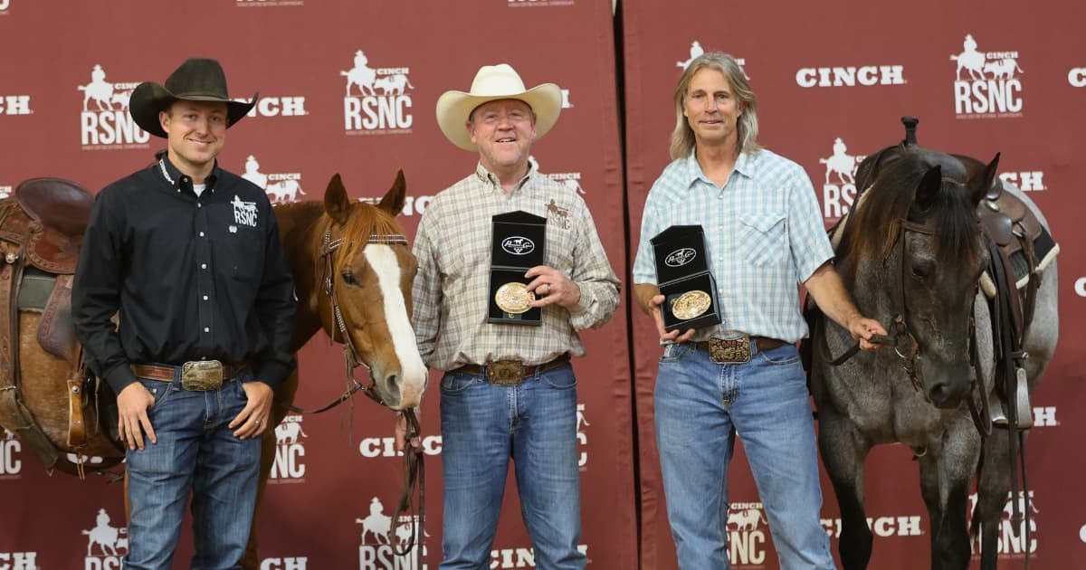 Two winners hold trophy buckles beside their horses and an RSNC official in front of a Cinch RSNC backdrop
