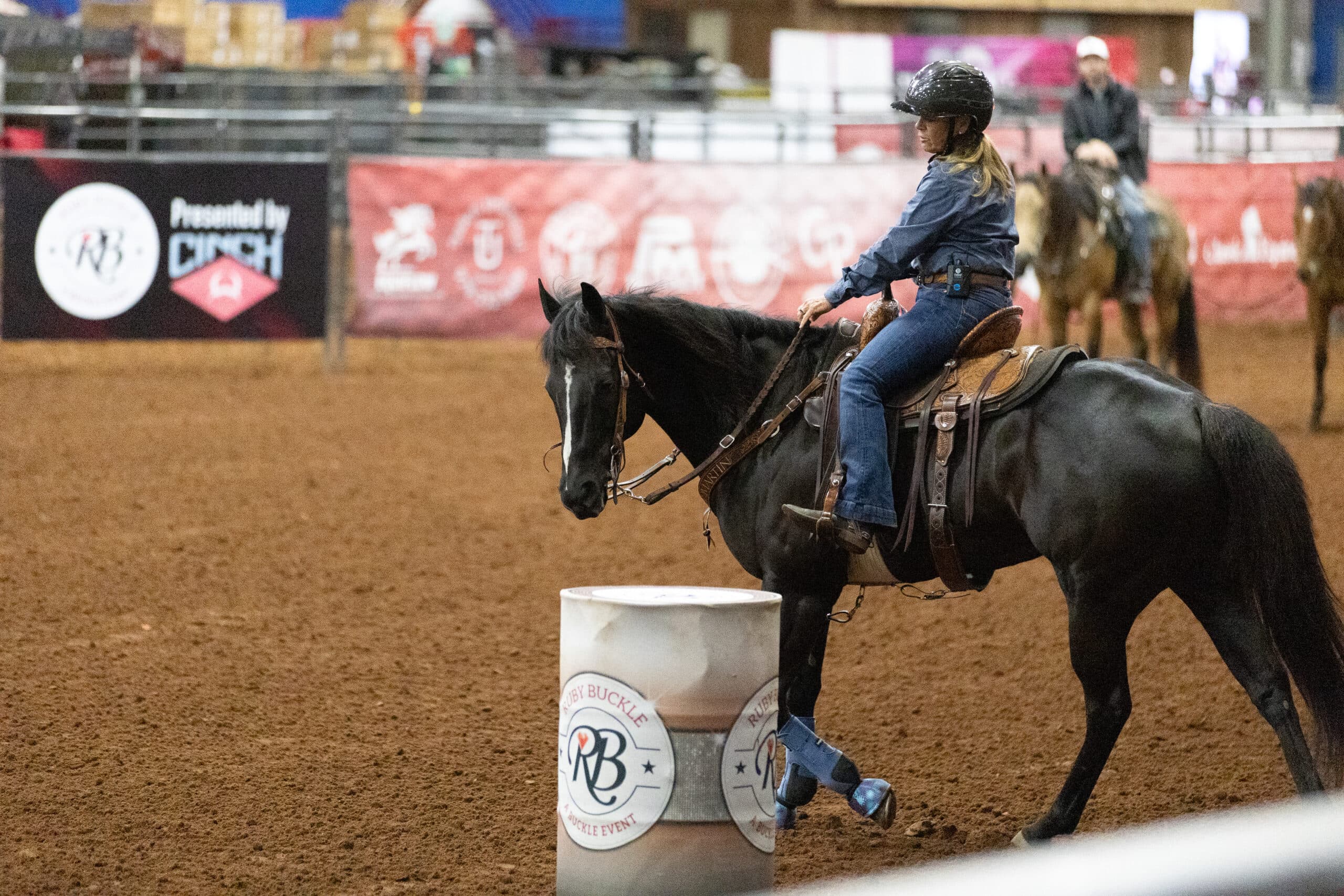 Rider in helmet and denim shirt guides a black horse in blue boots around a Ruby Buckle barrel in an indoor arena