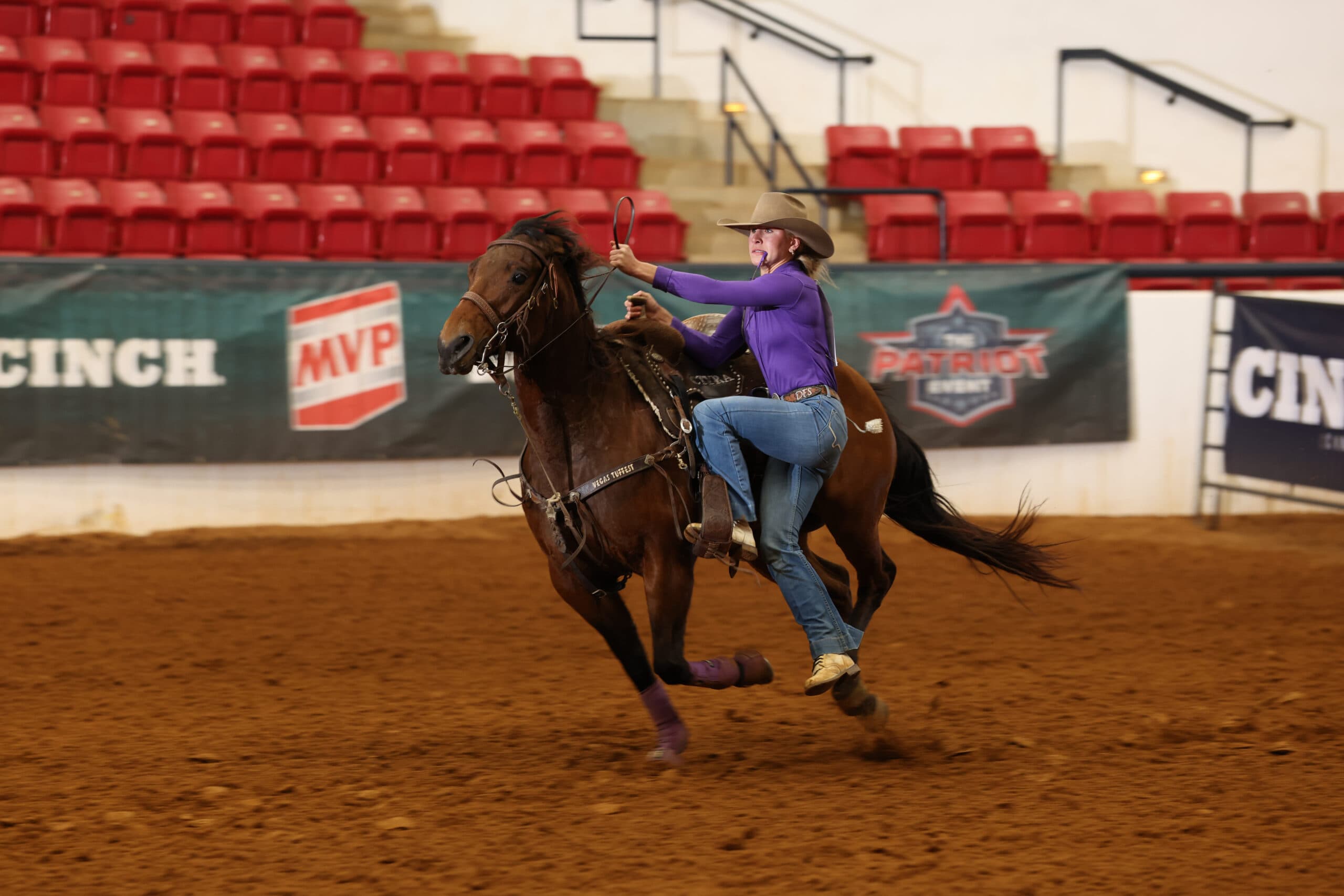 Barrel racer in a purple shirt and cowboy hat rides her bay horse hard around the arena past Cinch and MVP banners