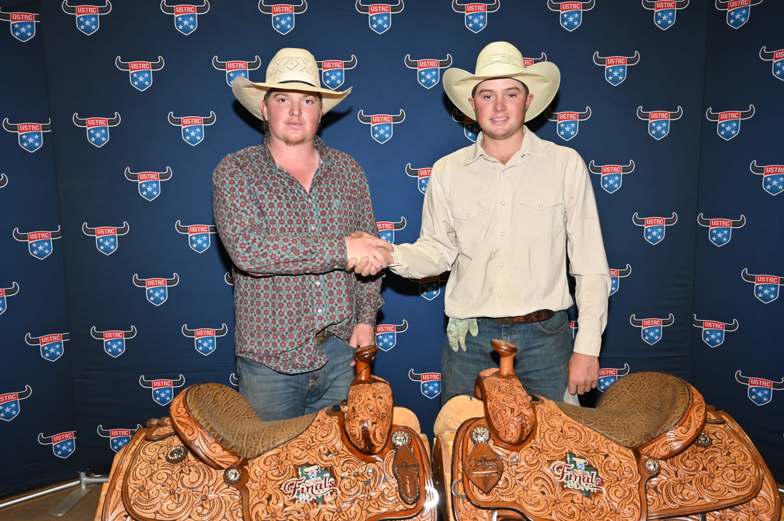 Two cowboys in Stetsons shake hands in front of a USTRC backdrop, standing beside two ornate western saddles