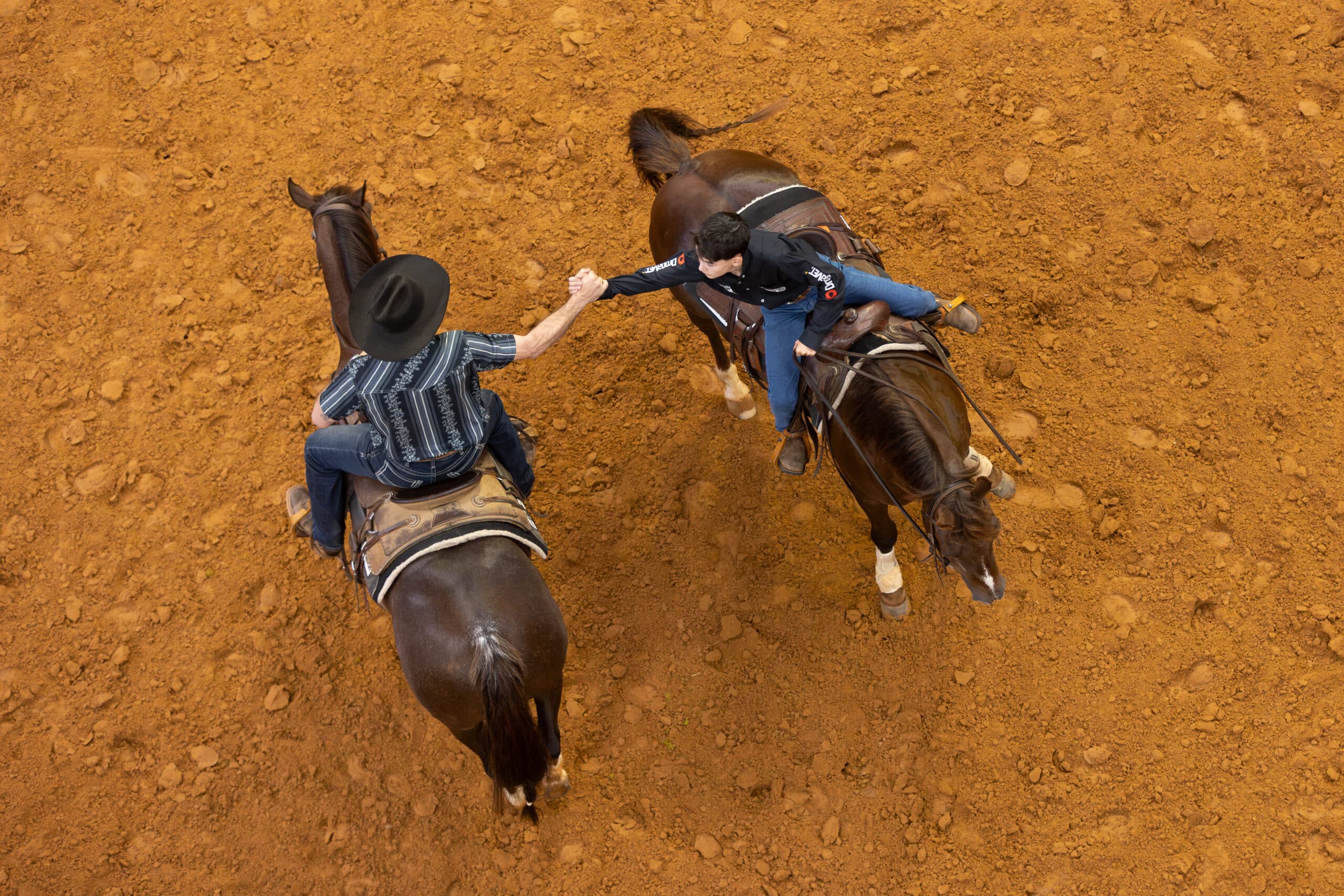 Overhead view of two mounted riders clasping hands in celebration on horseback in a red dirt arena