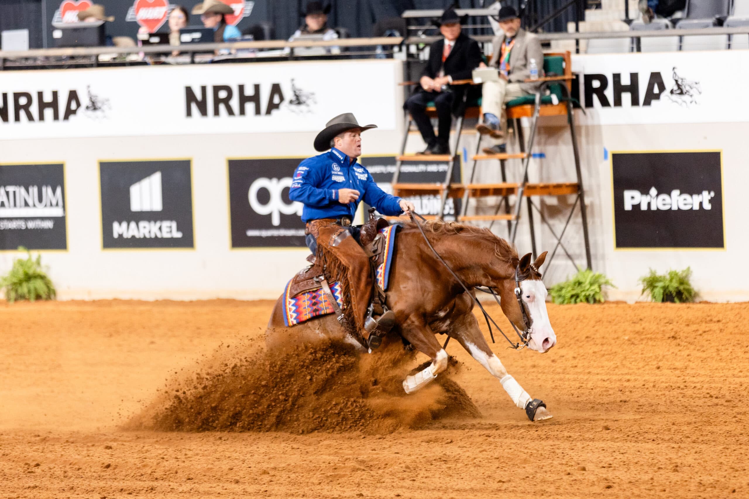 Reiner in blue shirt and chaps slides his chestnut bald-faced horse to a stop, spraying dirt in an NRHA arena