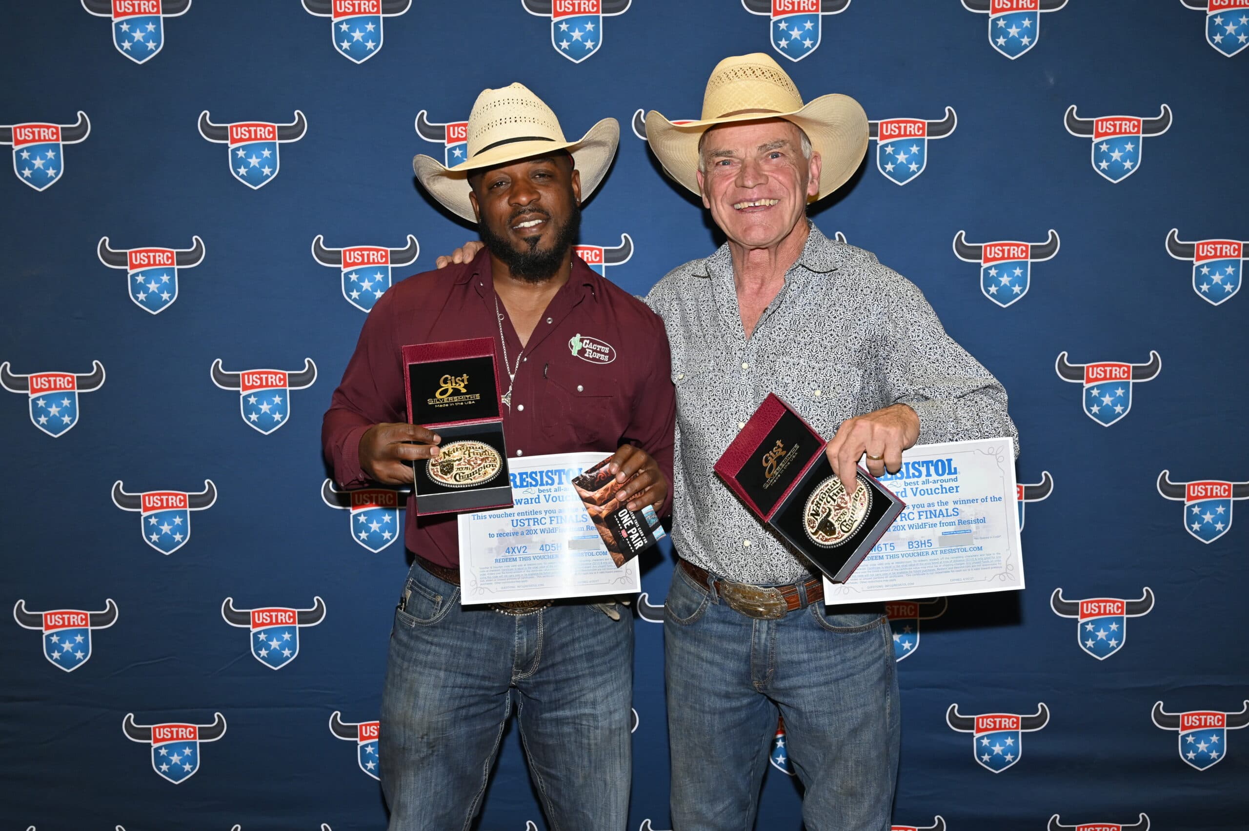 Two rodeo competitors in cowboy hats hold awards and certificates at a USTRC event backdrop