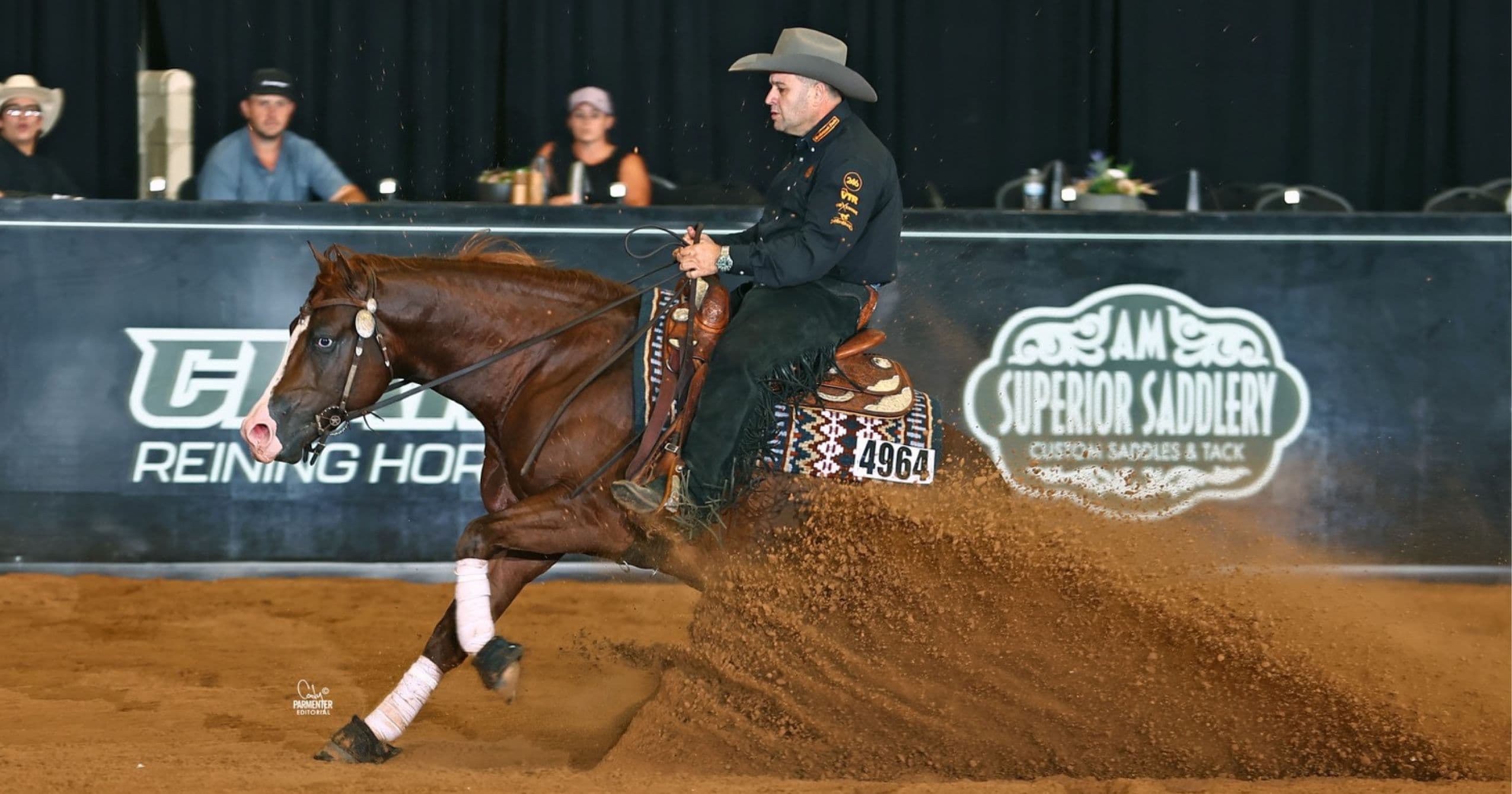 Reiner in black shirt and chaps slides his chestnut horse to a stop, spraying dirt in front of arena banners