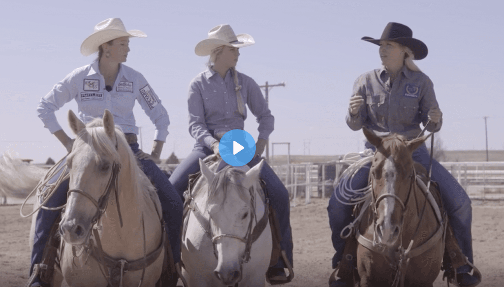 Three cowgirls in cowboy hats sit horseback with ropes coiled on their saddles, talking together in a dirt arena
