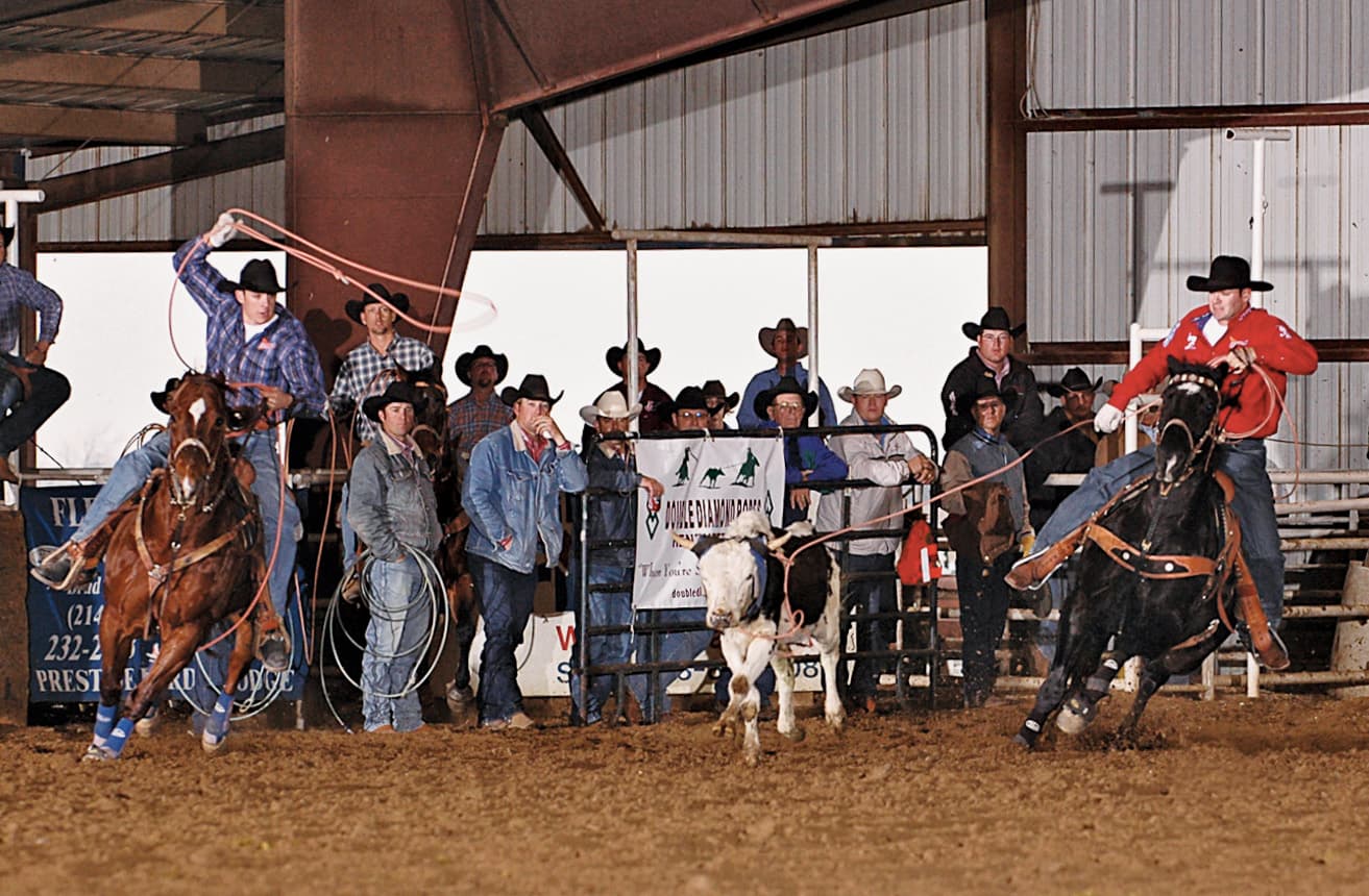 Two team ropers chase a steer across the arena as cowboys watch from the fence in an indoor rodeo