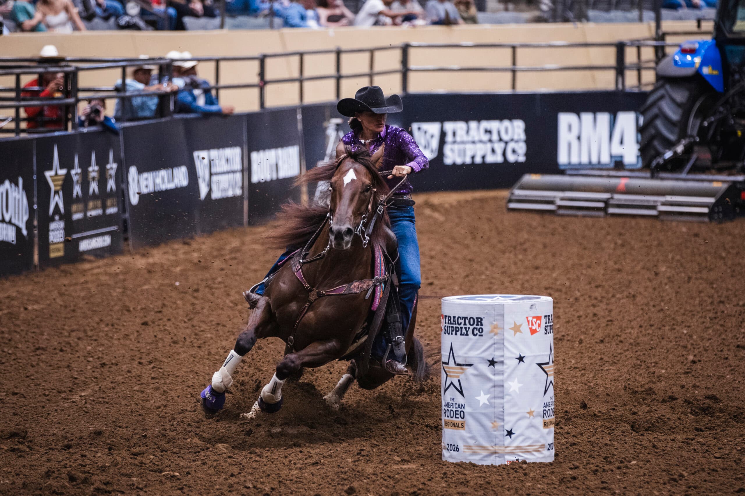 Barrel racer in a purple shirt and black cowboy hat rounds a Tractor Supply branded barrel on a sorrel horse