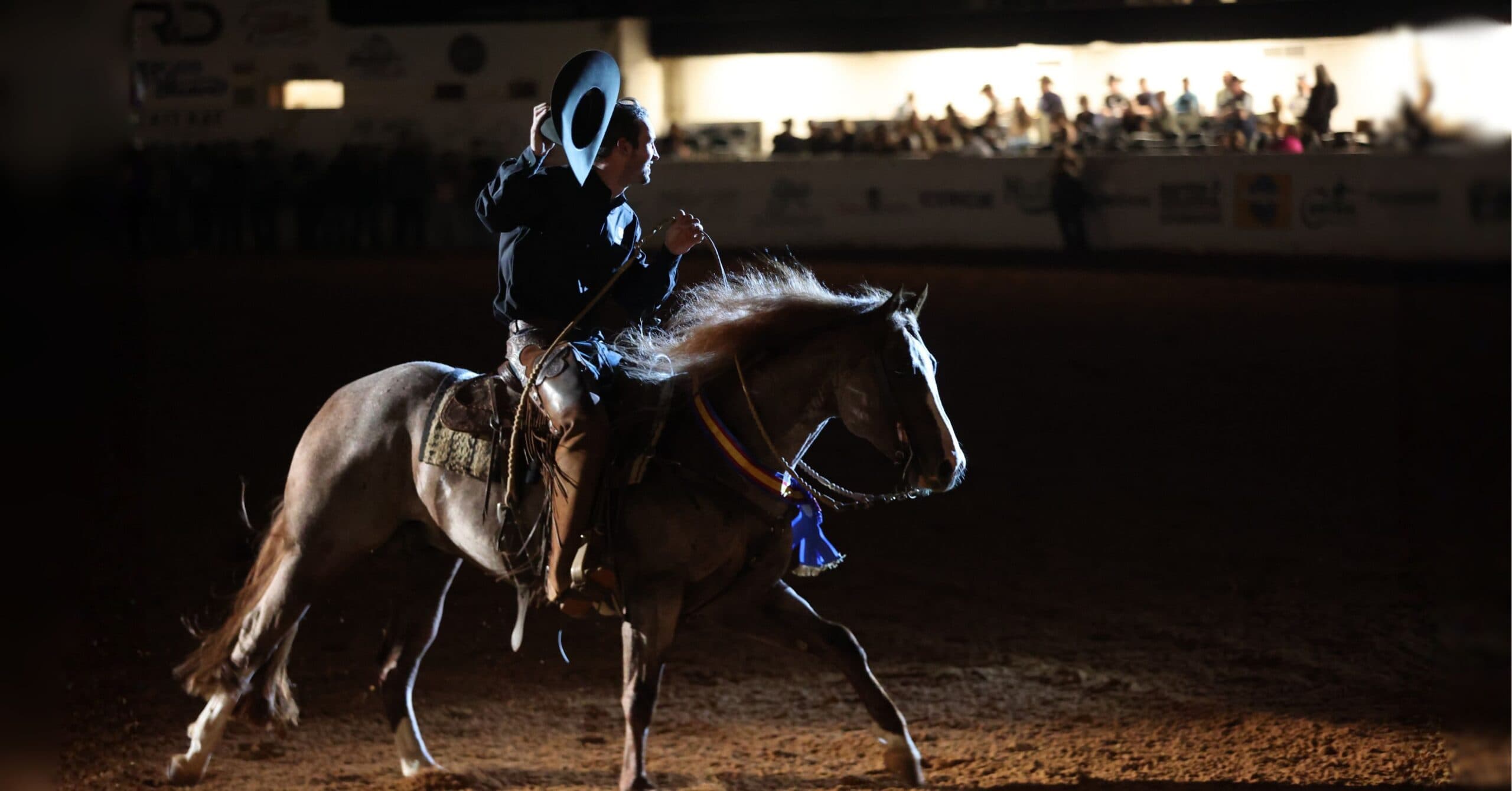 Cowboy tips his hat while riding a roan horse wearing a championship neck ribbon in a dimly lit arena