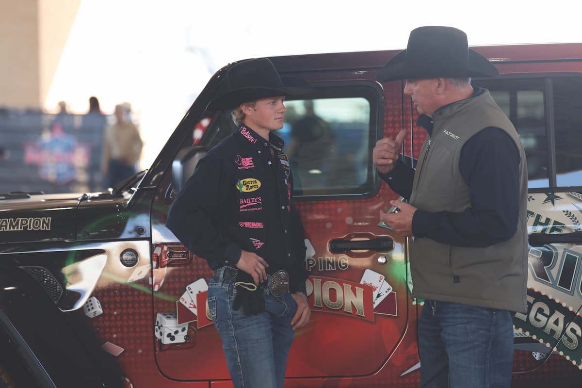 Young rodeo competitor in sponsored jacket converses with older man in cowboy hat beside red pickup truck at outdoor event