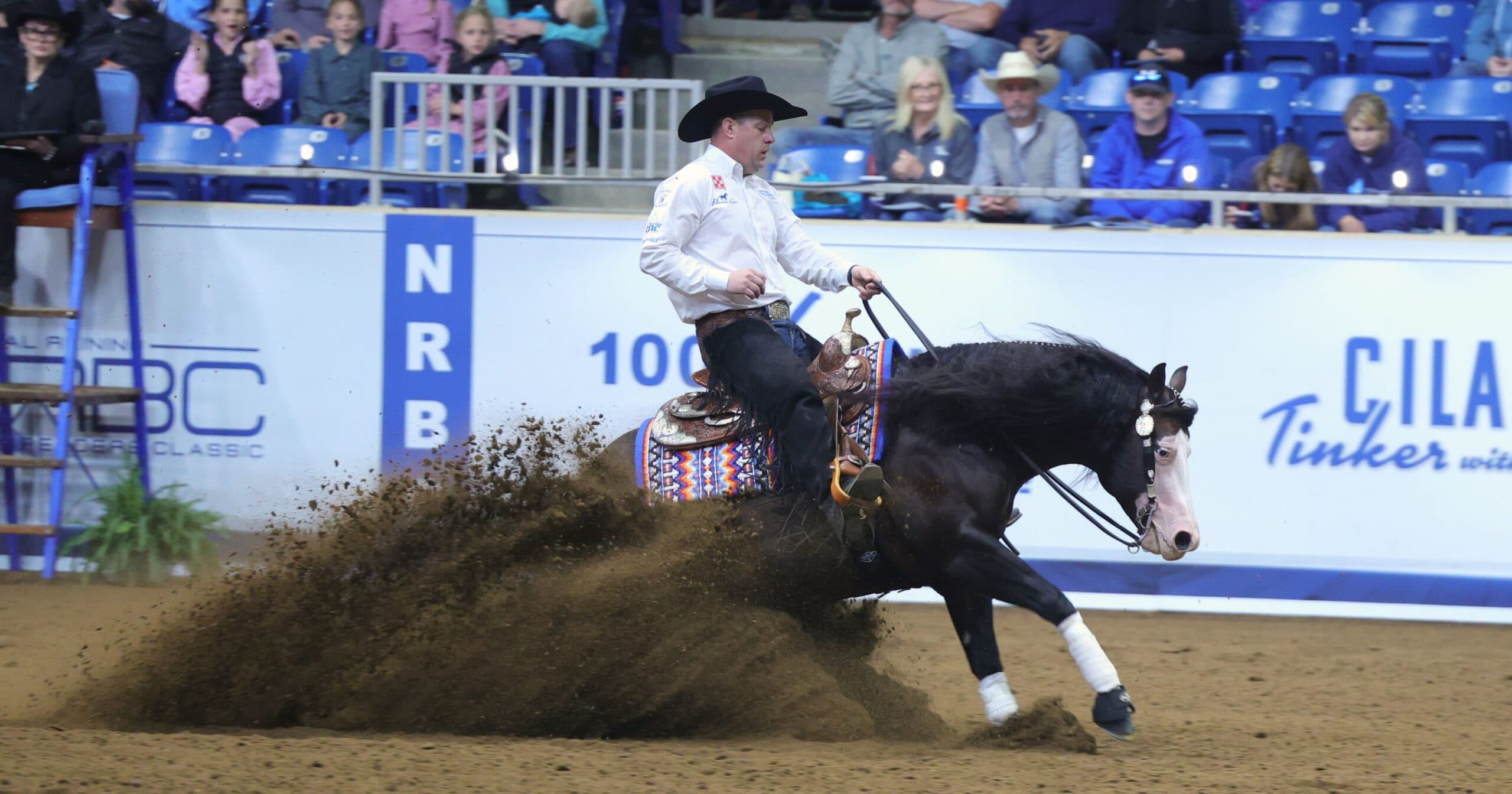 Reiner in black cowboy hat and white shirt slides his dark horse to a stop, dirt flying, before arena spectators