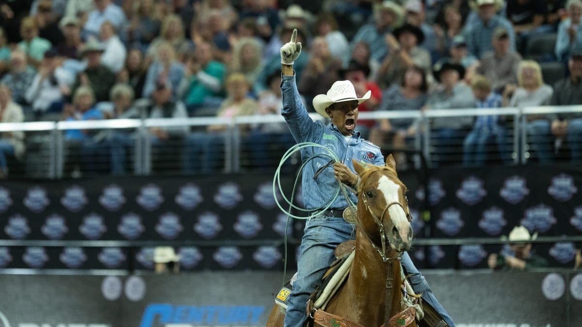 Rodeo competitor raises his fist in celebration while riding a brown horse and holding a lasso at a professional rodeo event with spectators in the stands