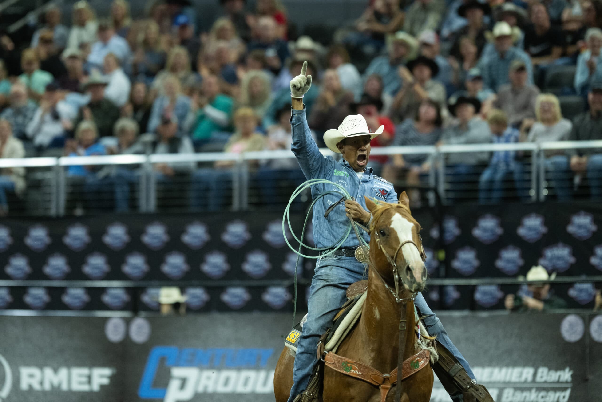 Rodeo competitor raises his fist in celebration while riding a brown horse and holding a lasso at a professional rodeo event with spectators in the stands