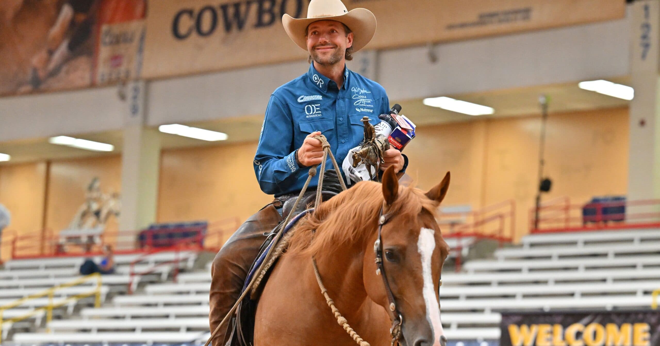 Smiling cowboy in a blue sponsor shirt and tan hat rides a chestnut horse through an indoor arena holding a bronze trophy and prizes