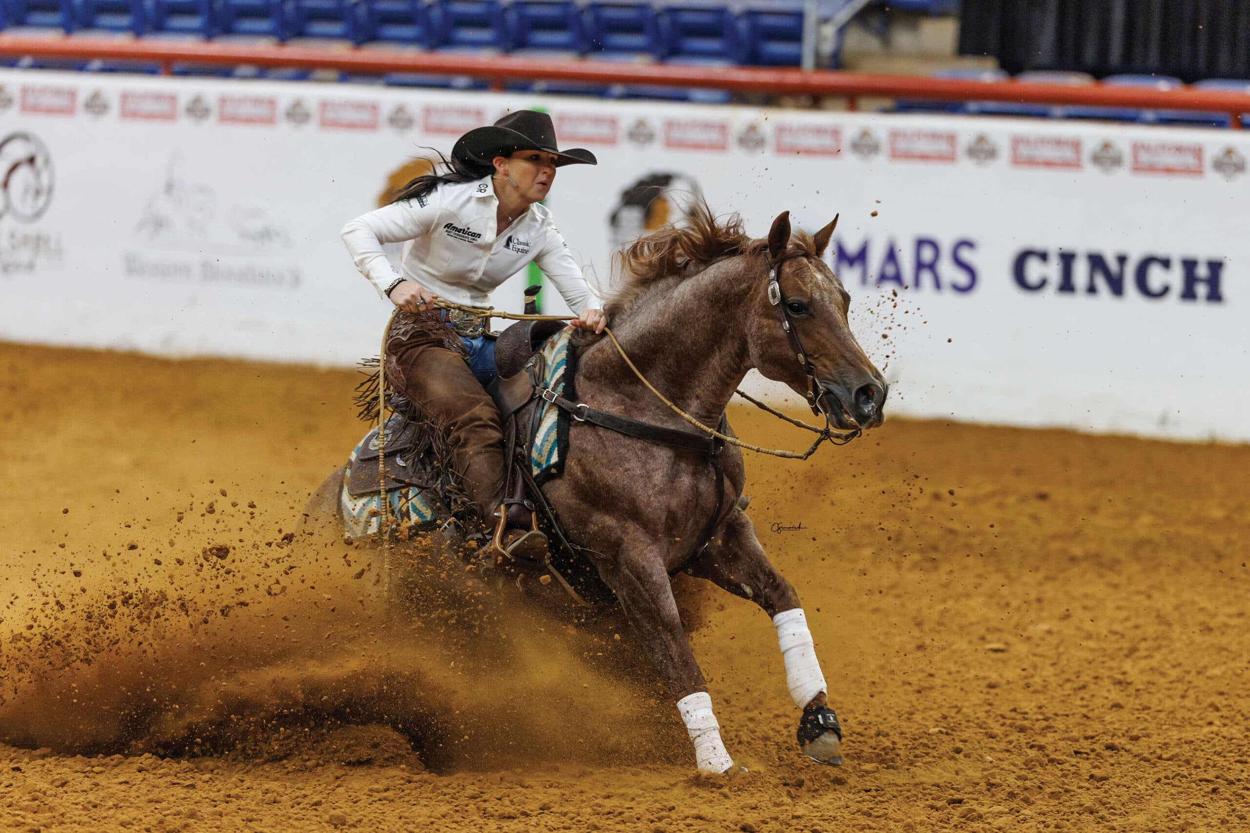 Barrel racer on gray horse rounds barrel at speed, kicking up dirt, wearing white shirt and black cowboy hat at indoor rodeo arena