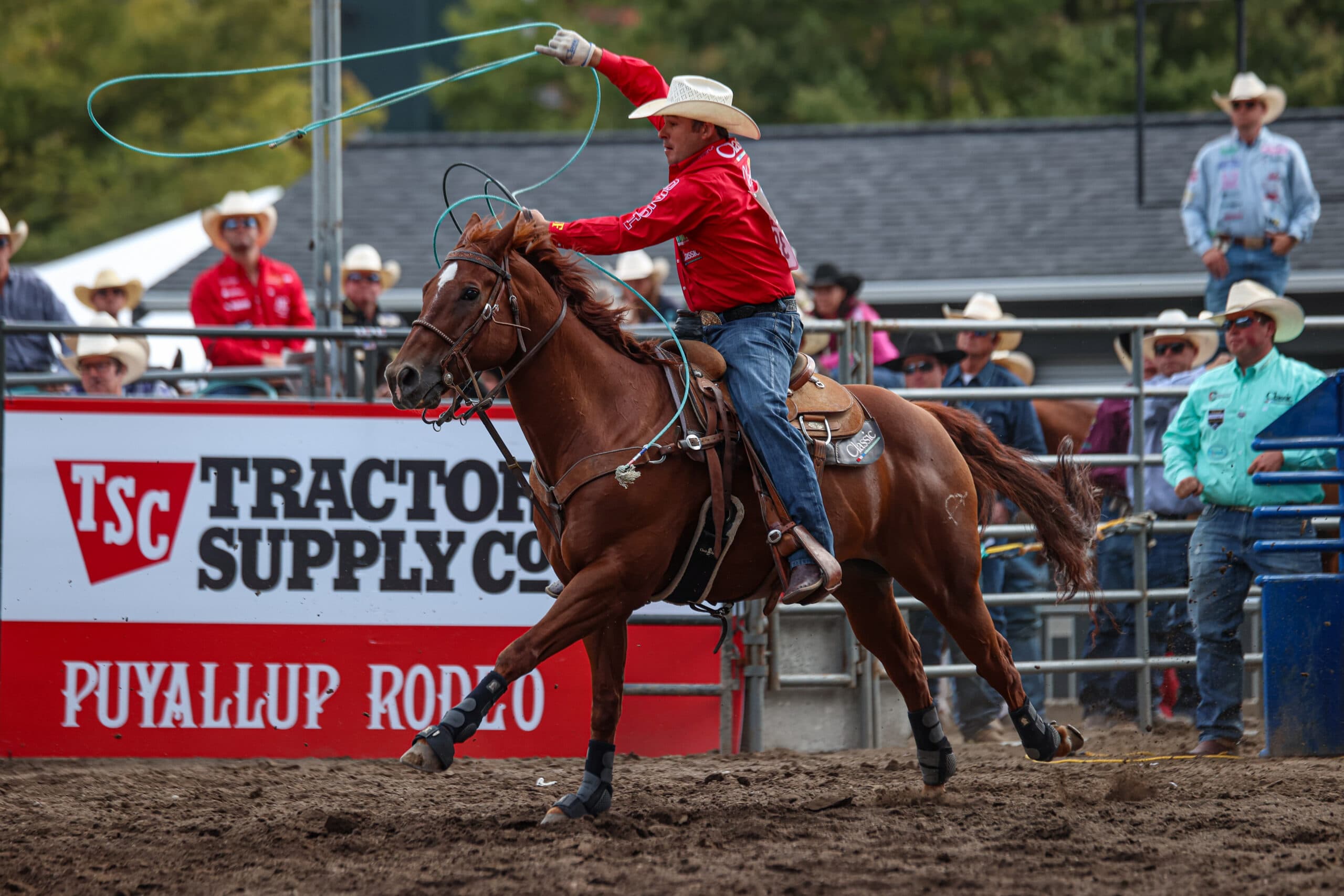 Roper in red shirt and straw hat swings a lasso while riding a brown horse at the Puyallup Rodeo, with spectators visible in the stands behind a Tractor Supply Co. banner