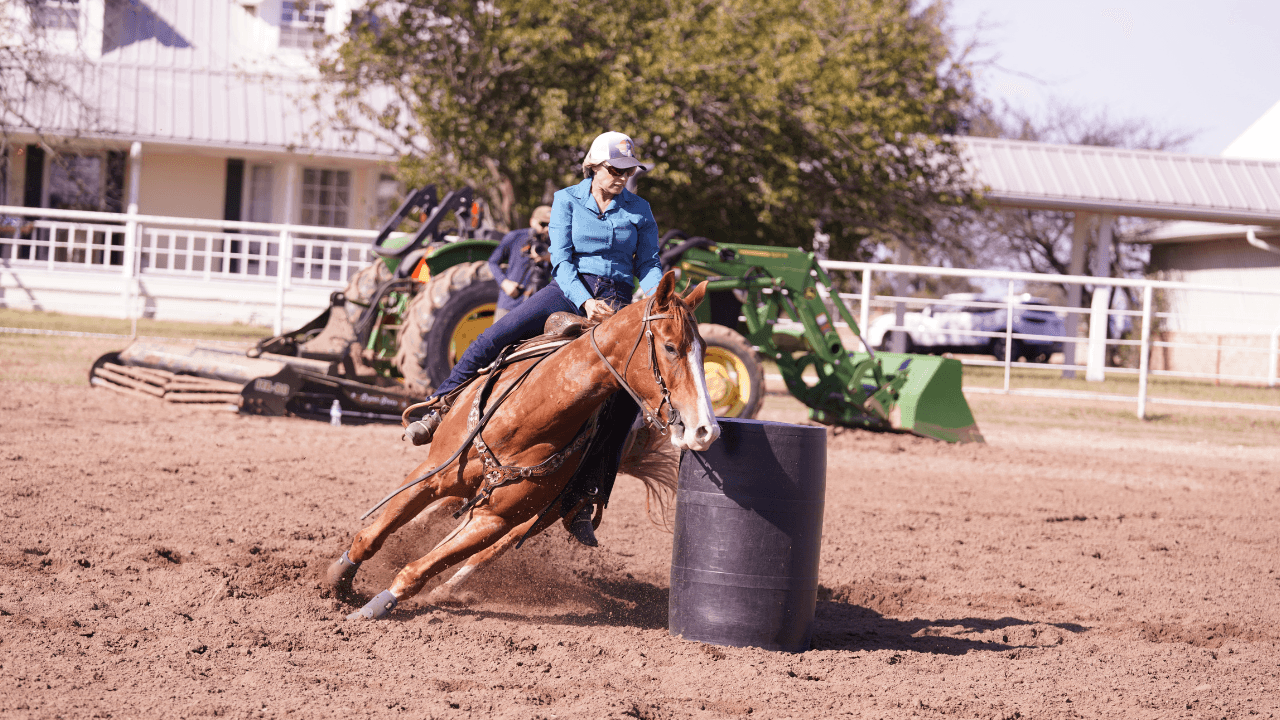 Rider in a teal shirt and ball cap turns a sorrel horse tightly around a black barrel, kicking up dirt in a practice arena