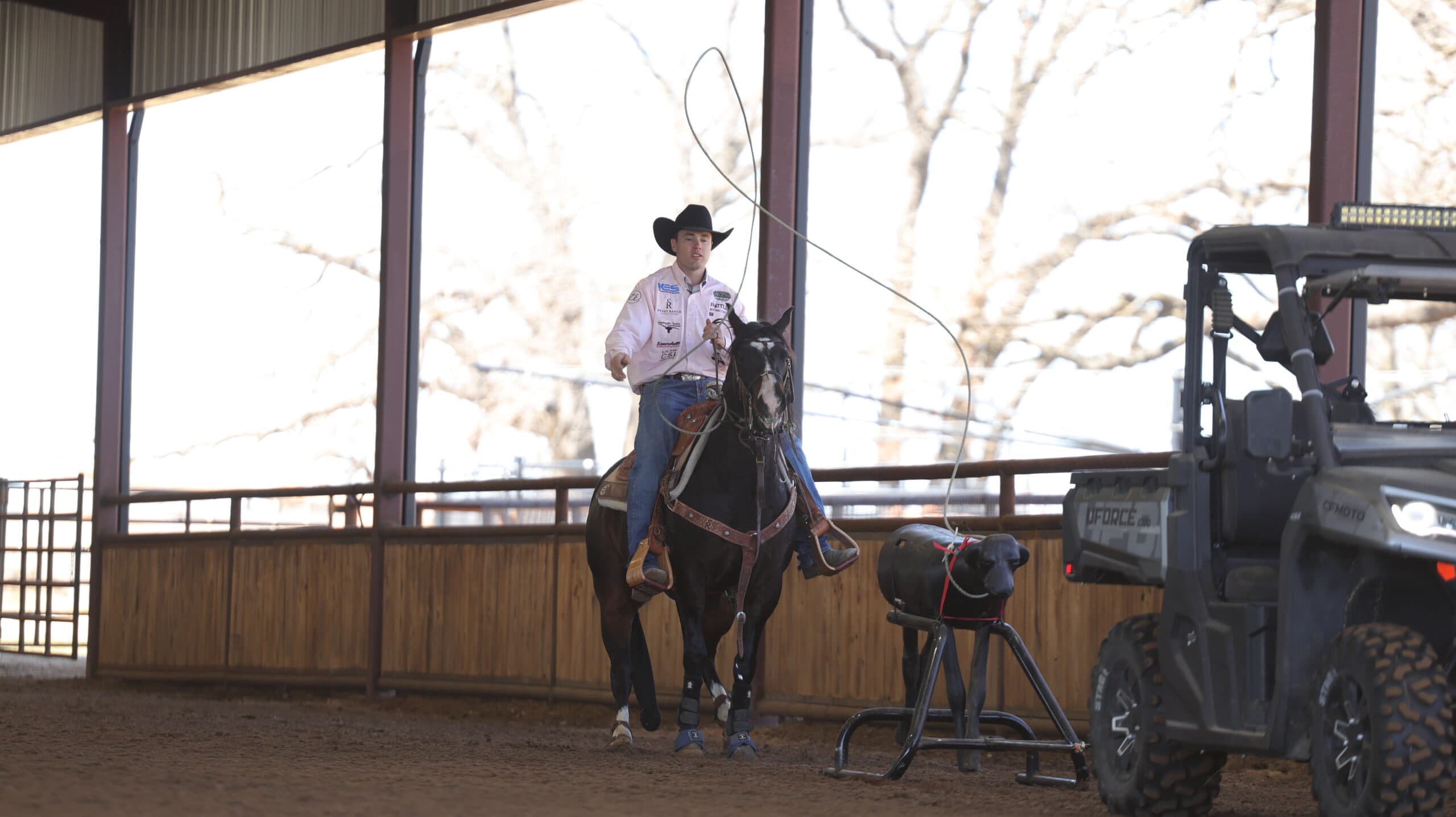 Cowboy in a black hat and pink shirt swings his rope aboard a dark horse while practicing on a roping dummy in a covered arena