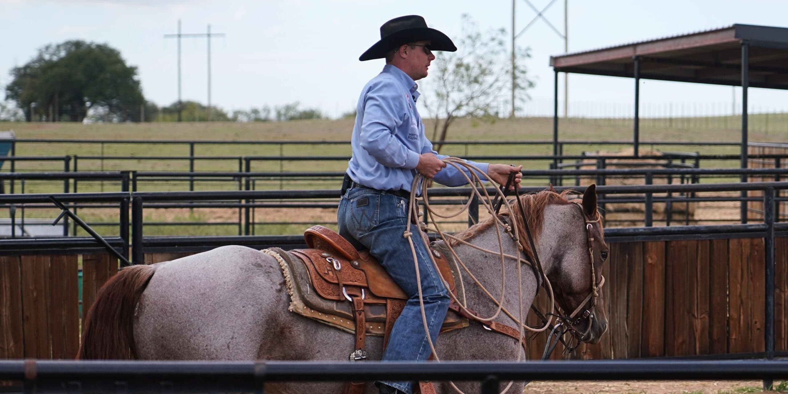 Cowboy in a black hat and blue shirt holds a coiled rope while riding a red roan horse in an outdoor pen