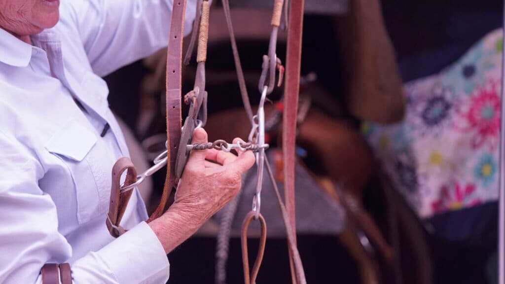 Weathered hands in a white button-down shirt inspect a leather bridle and curb chain bit hanging at a tack display