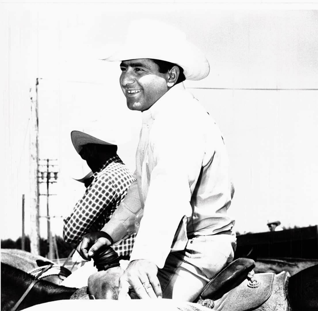 Smiling cowboy in white shirt and Stetson seated on horseback, wearing checkered sleeve guards and leather gloves