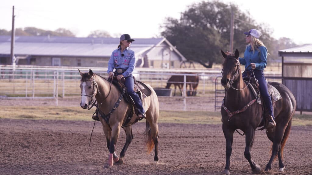 Two women in western shirts and ball caps ride horses side by side in a dirt arena, talking as they pass barns and pens