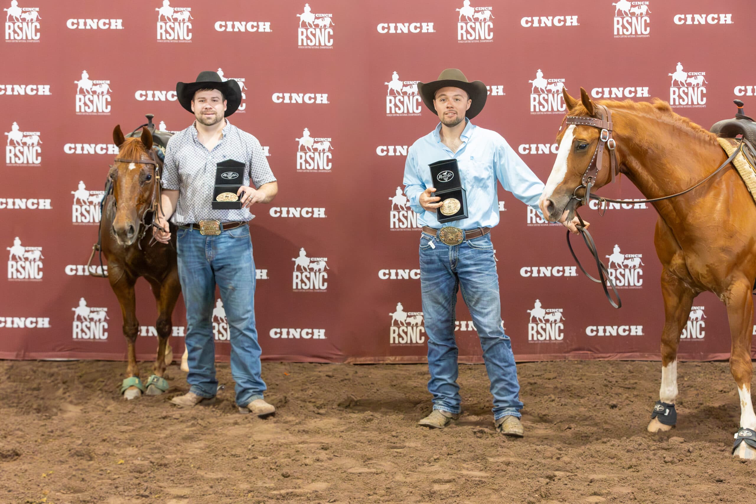 Two cowboys hold championship buckles in presentation boxes beside their sorrel horses in front of a Cinch RSNC backdrop