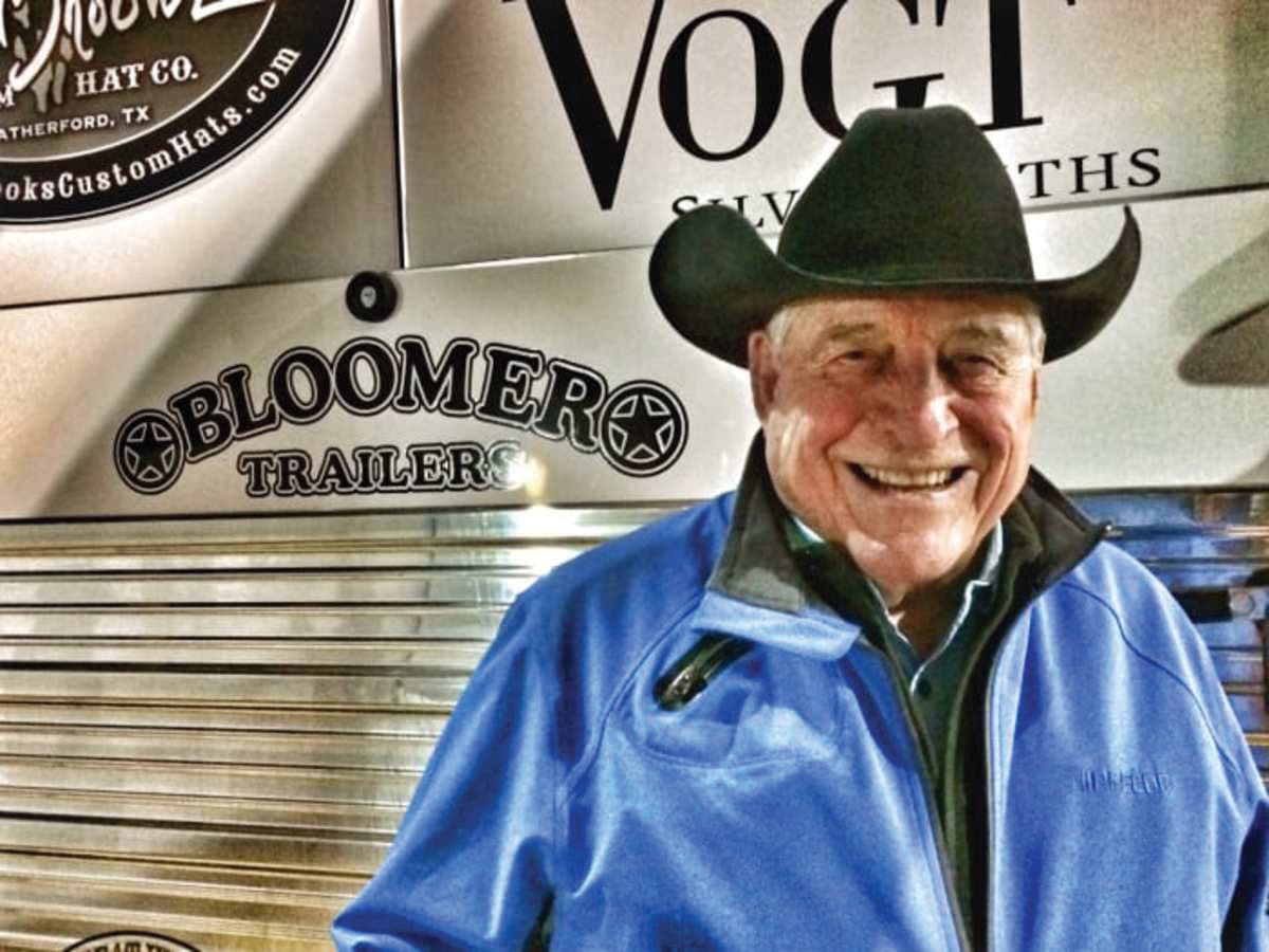 Smiling man wearing a green cowboy hat and blue jacket stands in front of a Bloomer Trailers sign