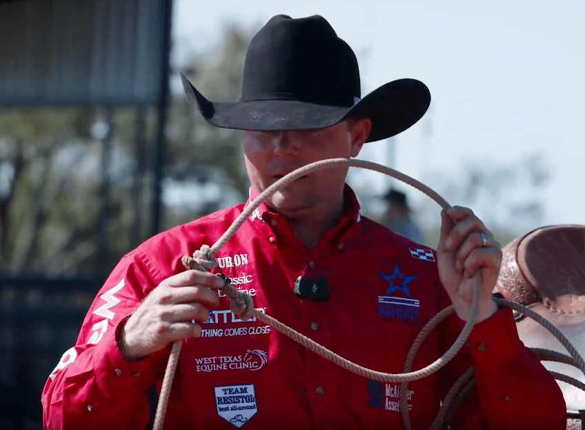 Roper in a black cowboy hat and red sponsor-covered shirt coils his rope while preparing to compete