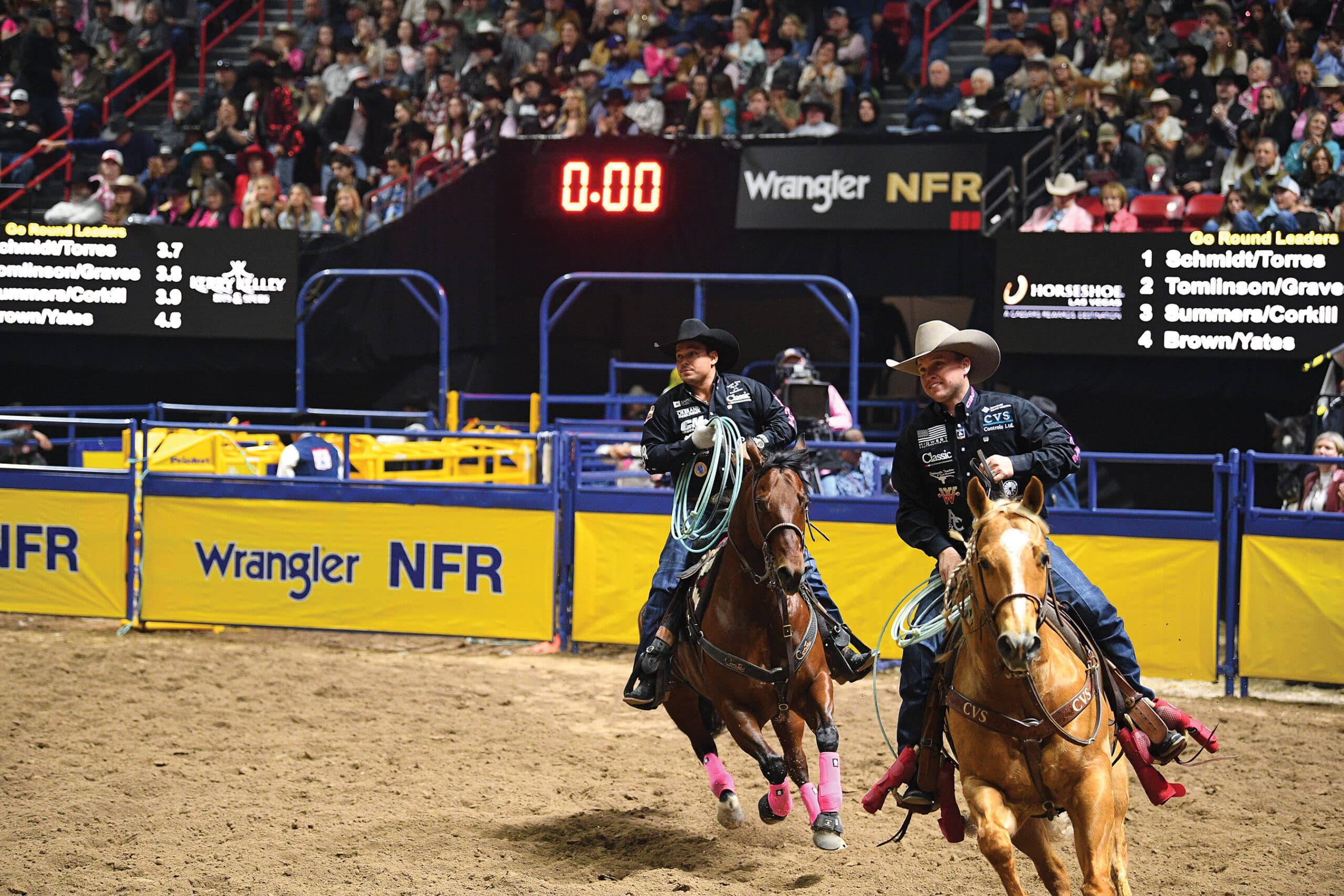 Two team ropers ride across the arena past the Wrangler NFR banner as the clock reads 0.00 in a packed Las Vegas crowd