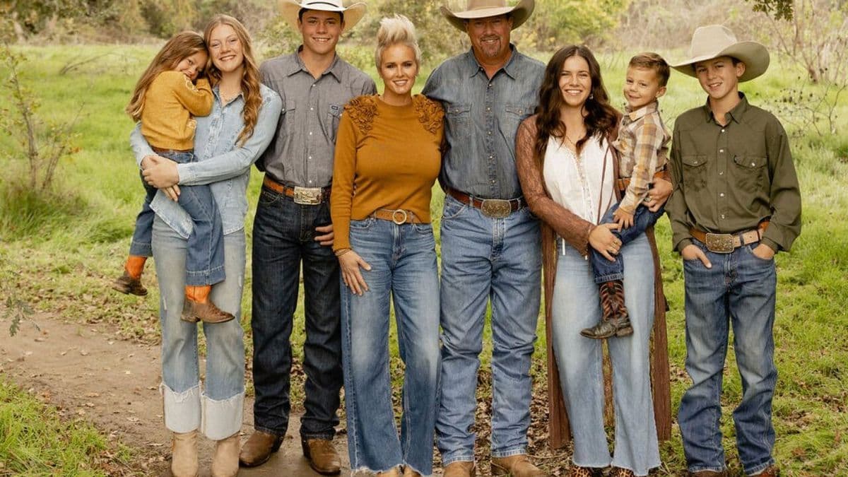 Family of eight in cowboy hats, denim and boots stands smiling side by side on a grassy dirt path outdoors