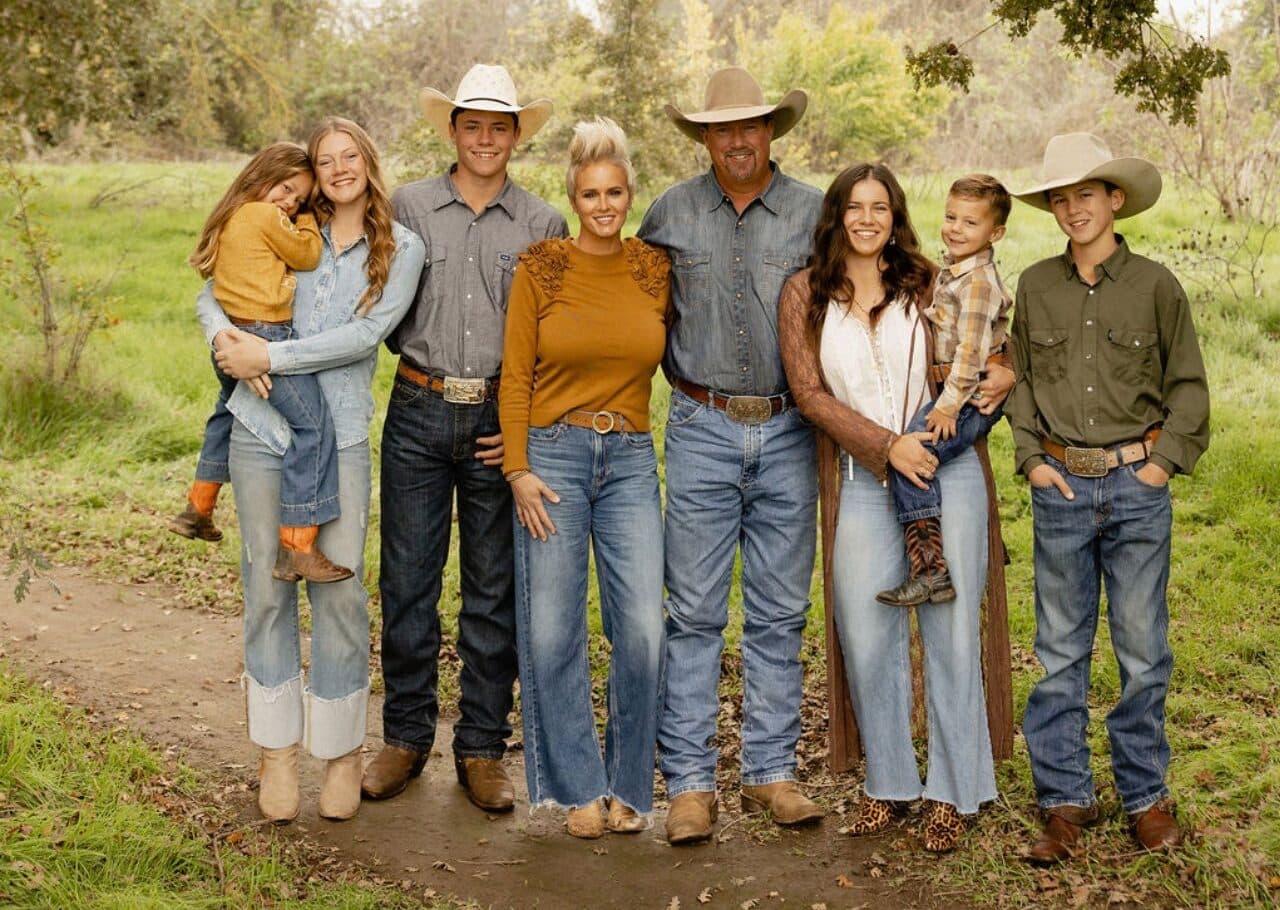 Family of eight in cowboy hats, denim and boots stands smiling side by side on a grassy dirt path outdoors