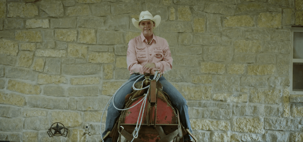Smiling cowgirl in a white hat and pink sponsor shirt sits mounted with rope coils in hand against a stone wall