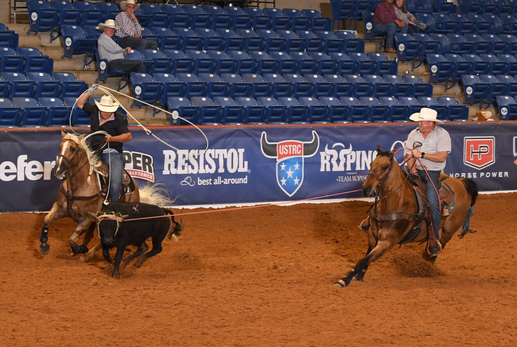 Team ropers compete during a USTRC sanctioned event, with one roper swinging a lasso toward a steer while a header positions his horse
