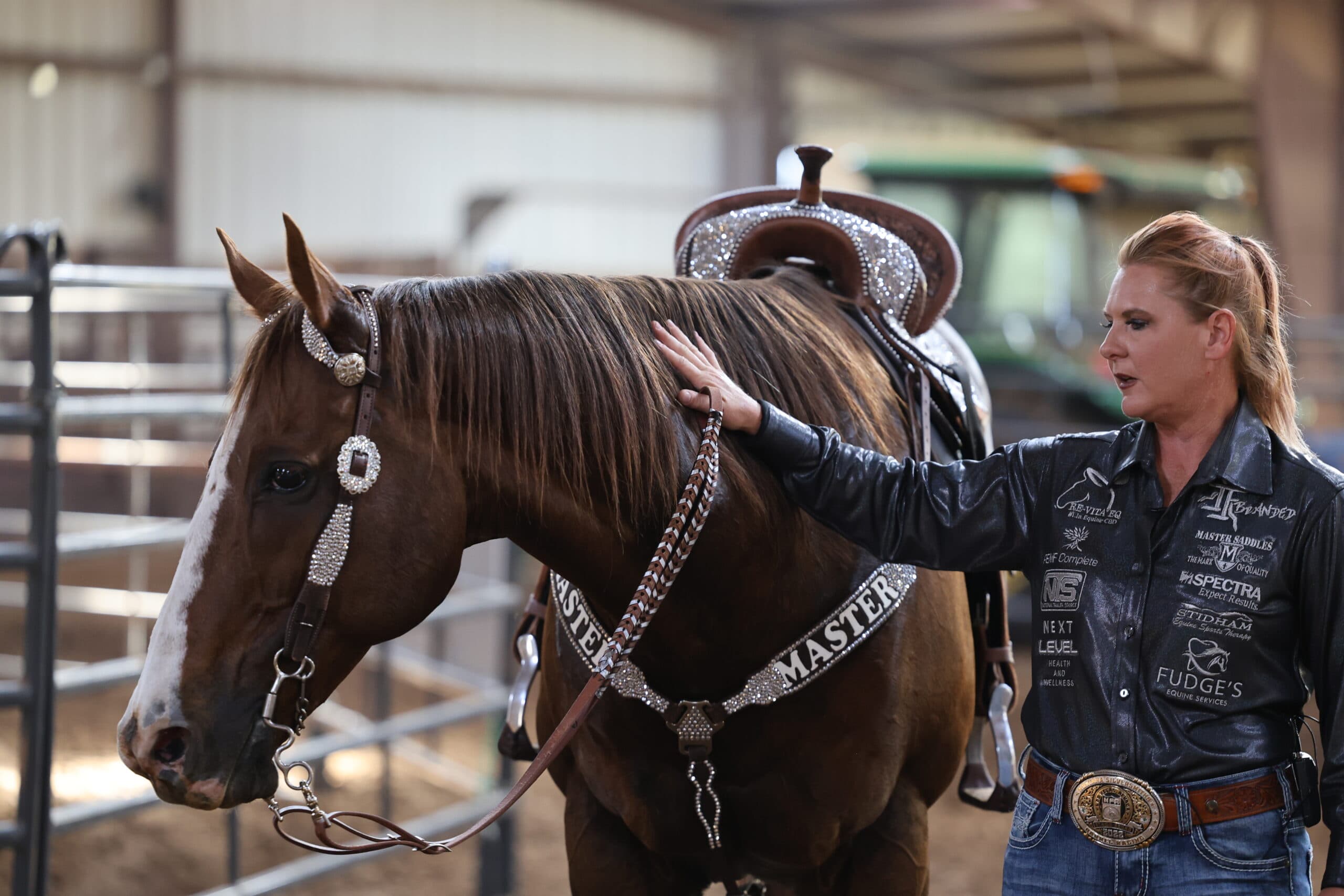 Woman in a sponsor-covered black shirt pats the neck of a sorrel horse wearing a rhinestone headstall and breast collar