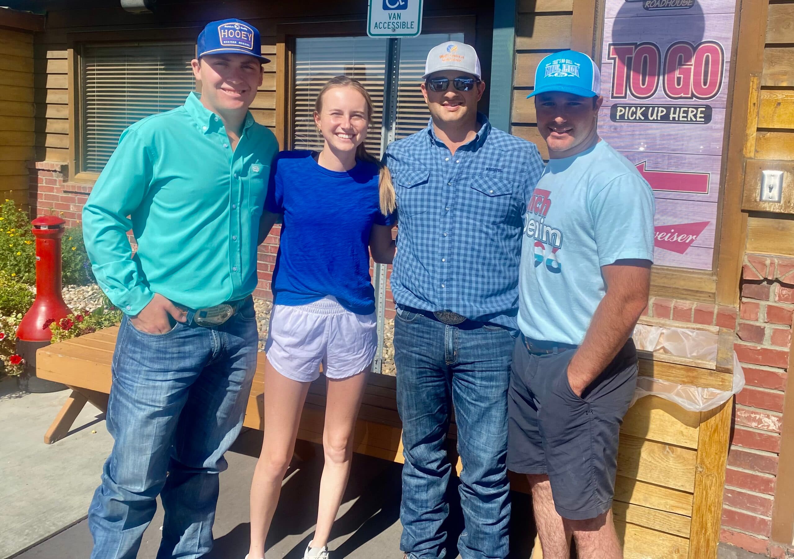 Three men in ball caps and a smiling woman pose arm in arm outside a roadhouse restaurant's to-go entrance