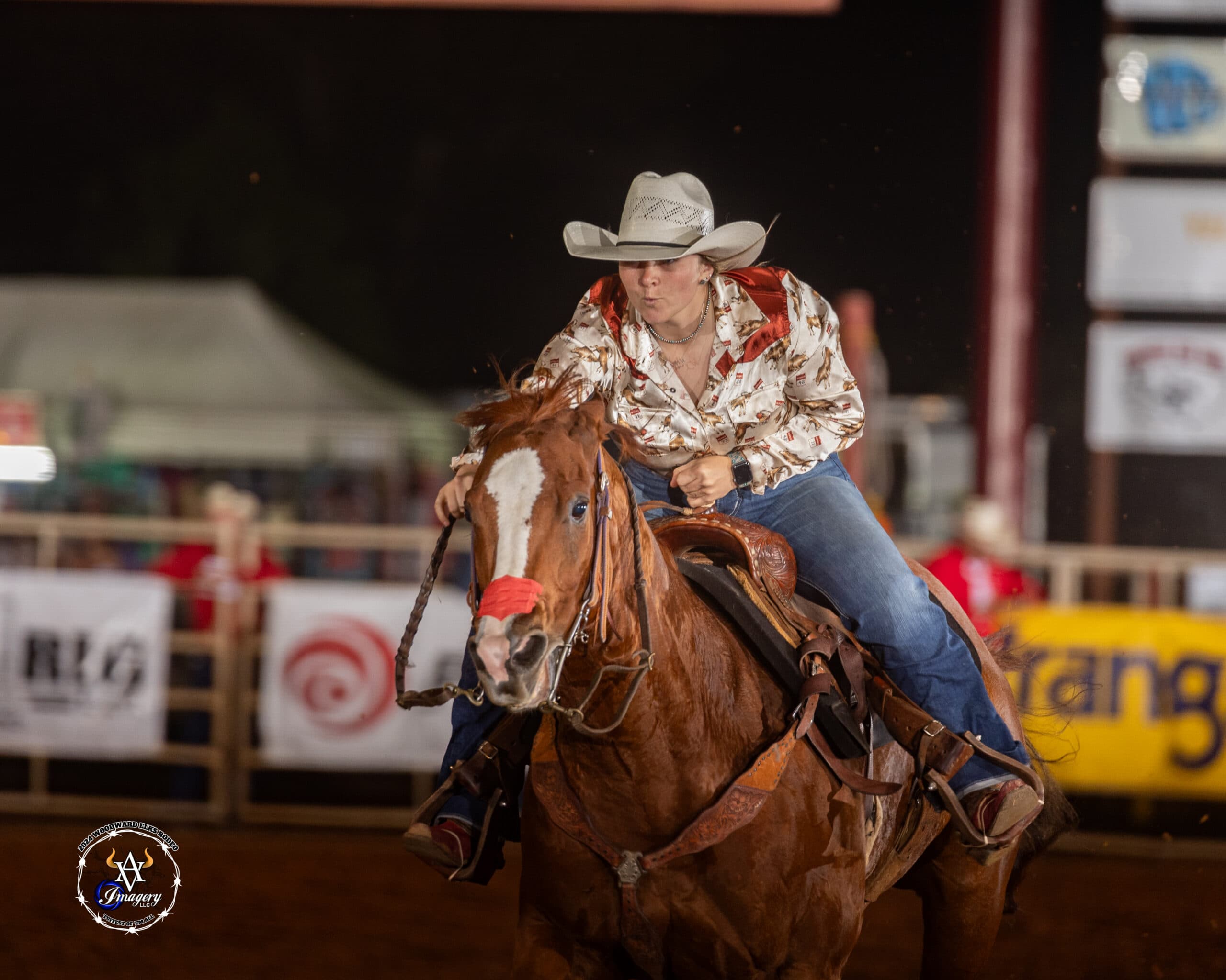 Barrel racer in a straw cowboy hat and printed western shirt leans forward as her sorrel horse rounds a turn under arena lights