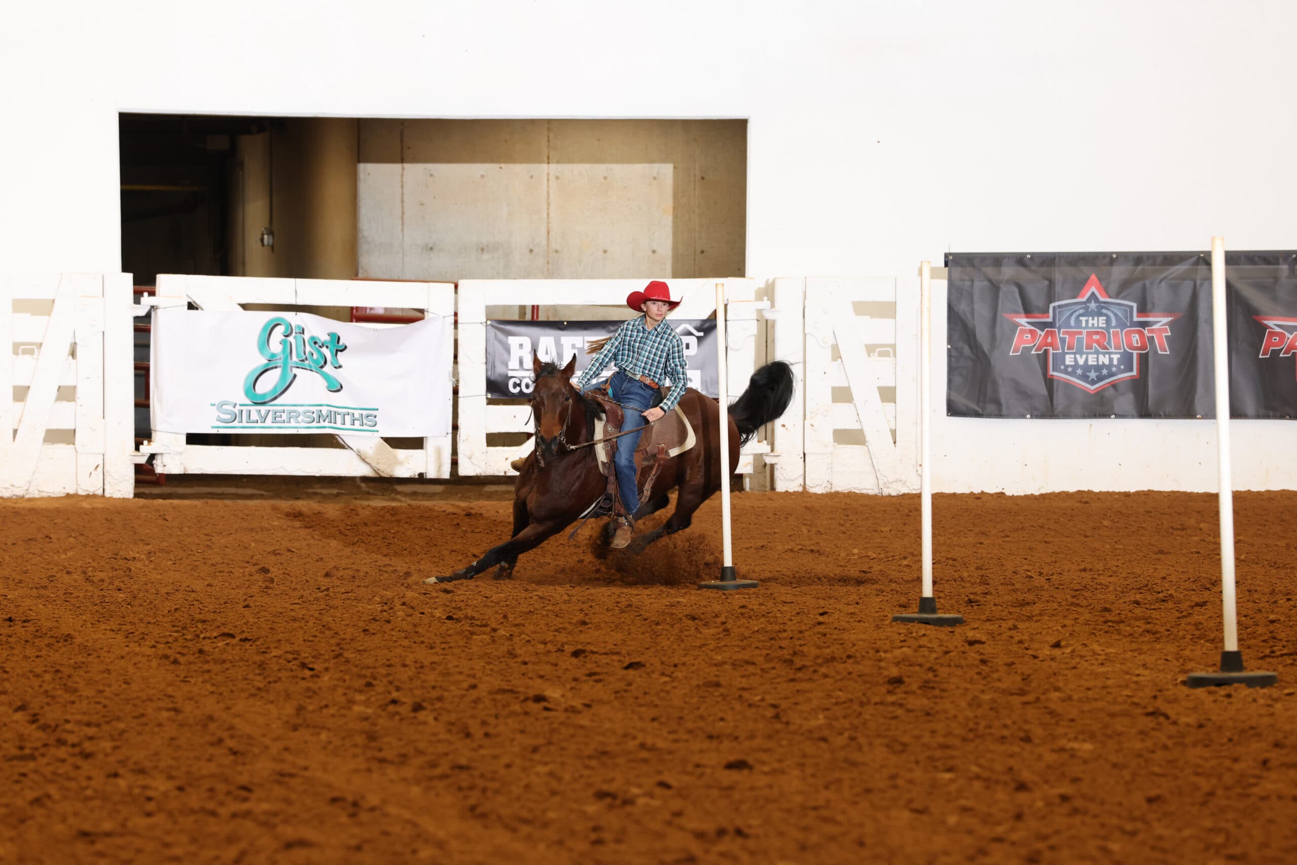 Young rider in a red cowboy hat and plaid shirt weaves her bay horse through pole bending poles in a dirt arena