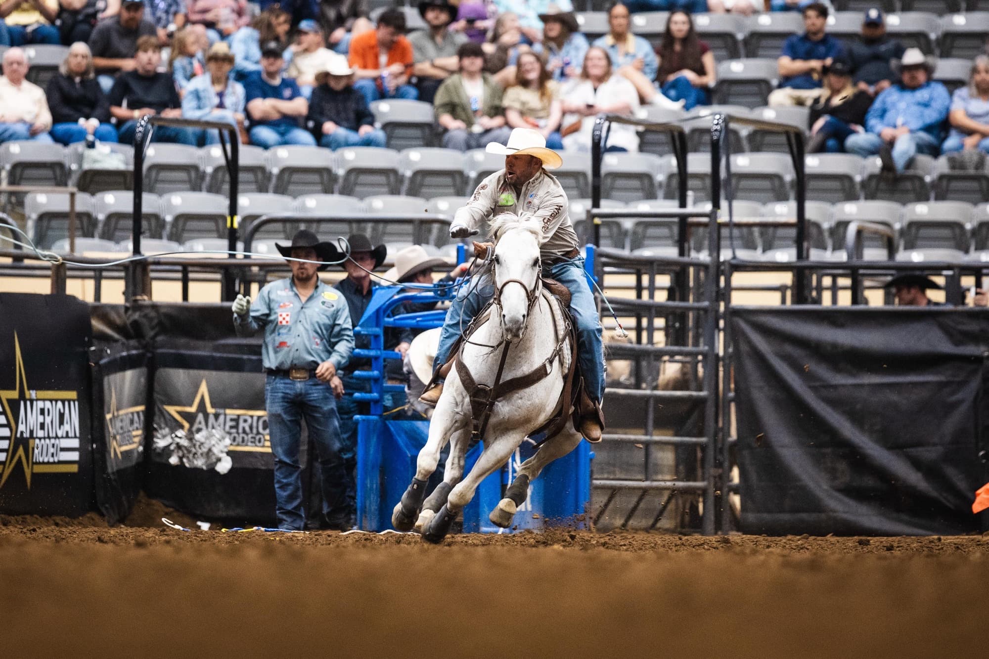 Cowboy on white horse galloping past blue barrier gates at rodeo arena with spectators in stands behind black fencing