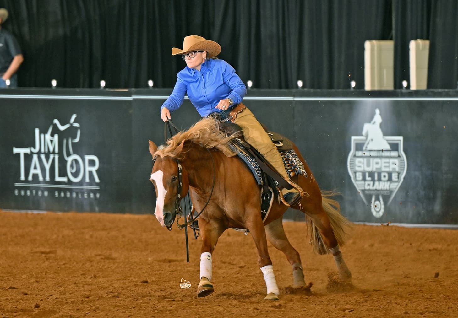 Reining rider in a blue shirt and tan cowboy hat sliding to a stop on a chestnut horse in a dirt arena