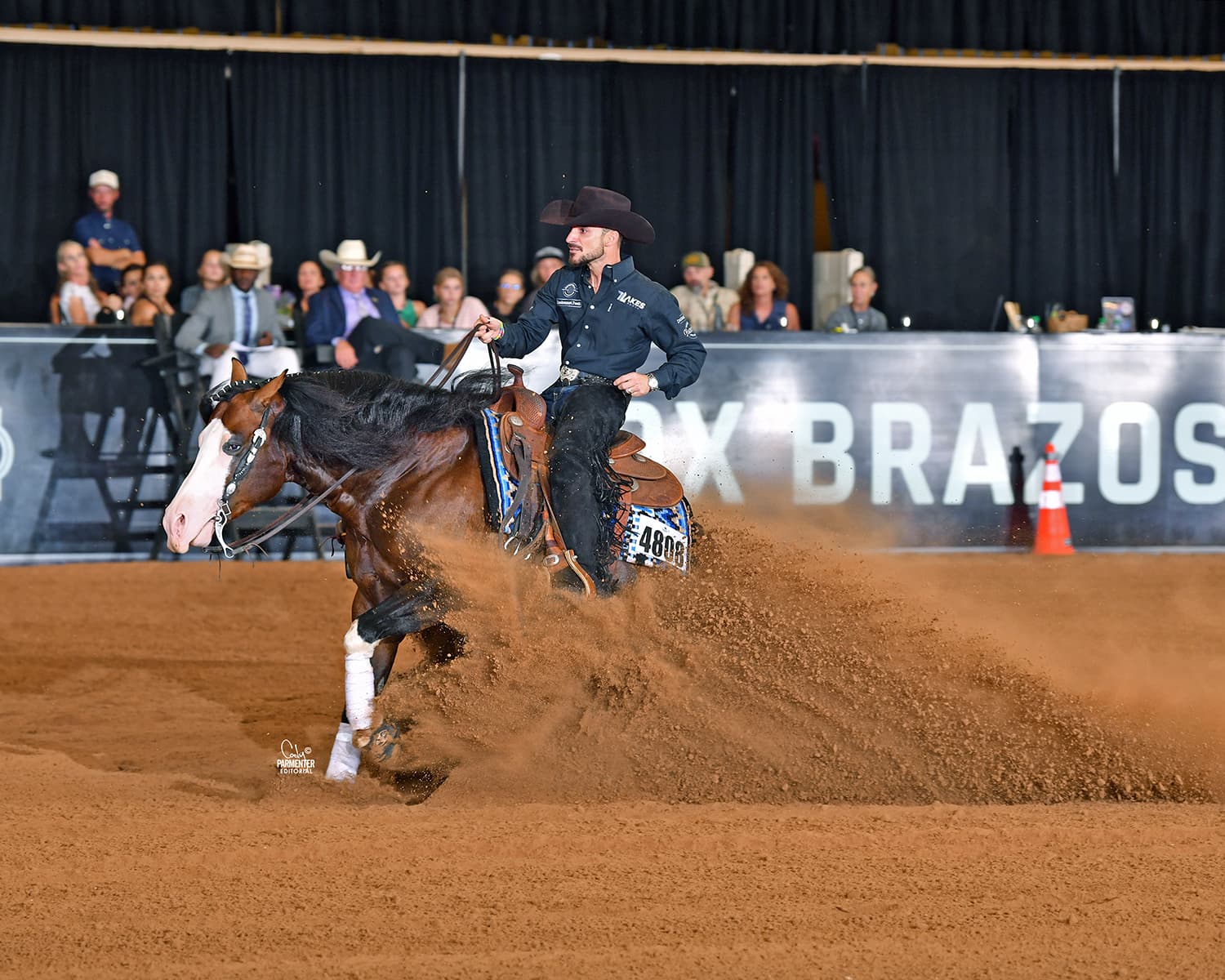 Reiner in a black cowboy hat slides his bay horse to a stop, spraying dirt in front of an indoor arena crowd