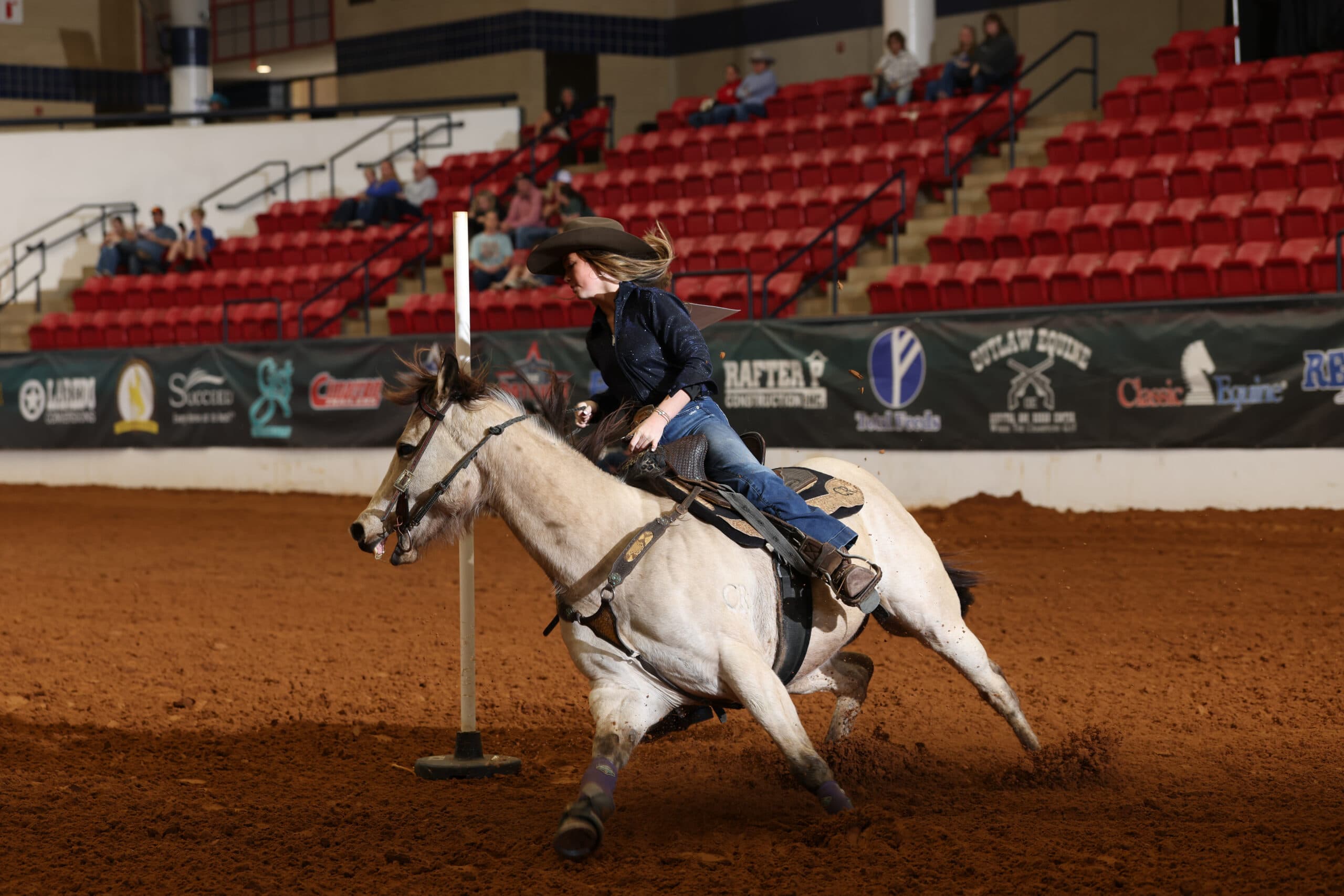 Cowgirl in a dark shirt and felt hat turns her buckskin horse sharply around a pole in an indoor arena during pole bending
